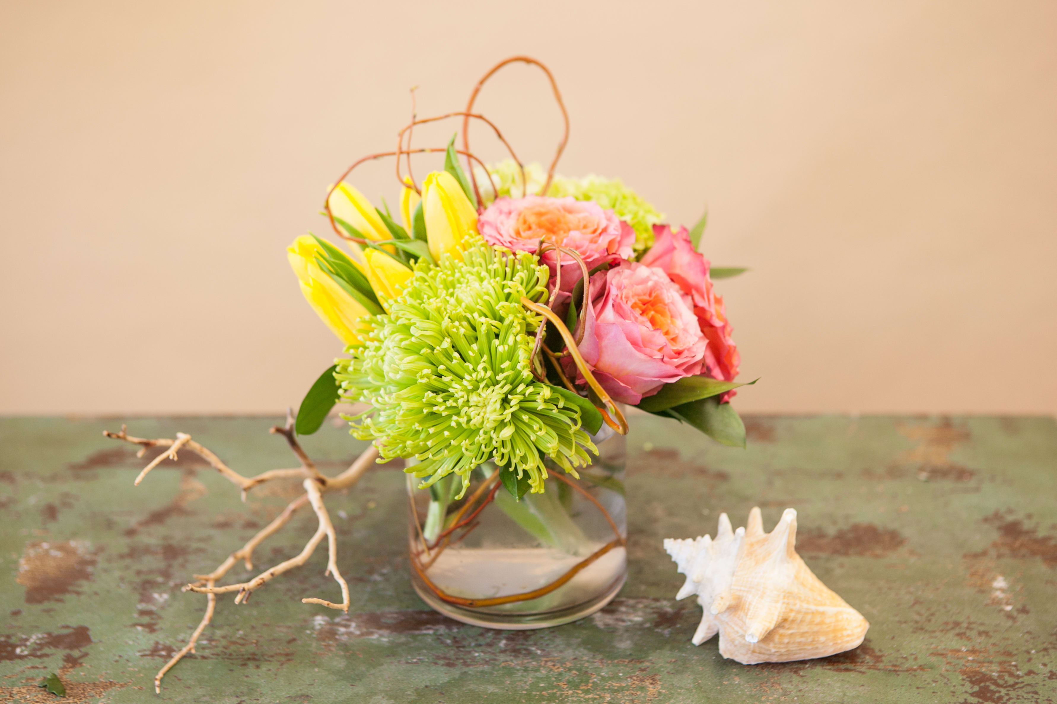 Pink and yellow flowers arranged in a clear glass vase with a seashell beside it