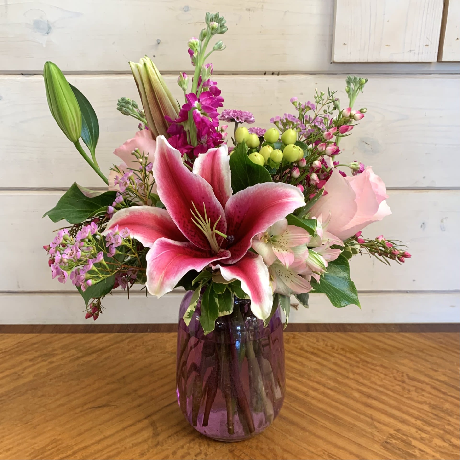 Sweet Nothings - Pale pink roses and stargazer lilies with fragrant magenta stock, green hypericum berries, and lavender button chrysanthemums arranged in a lavender tulip vase with pink alstroemeria and seasonal filler flower. Approximately 11" wide by 13" high.