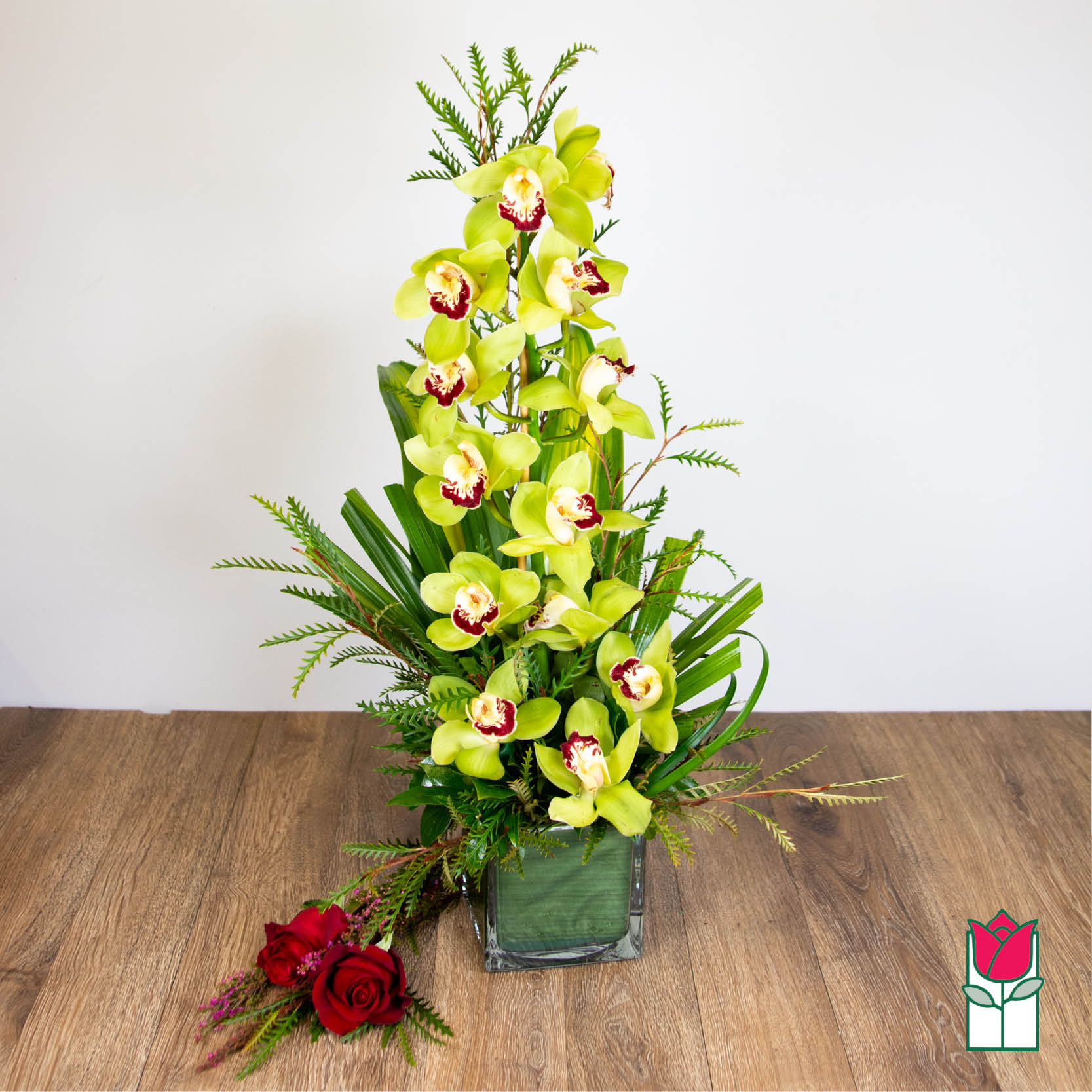 Tall green cymbidium orchid arrangement in a leaf-lined glass cube vase with two red roses beside it