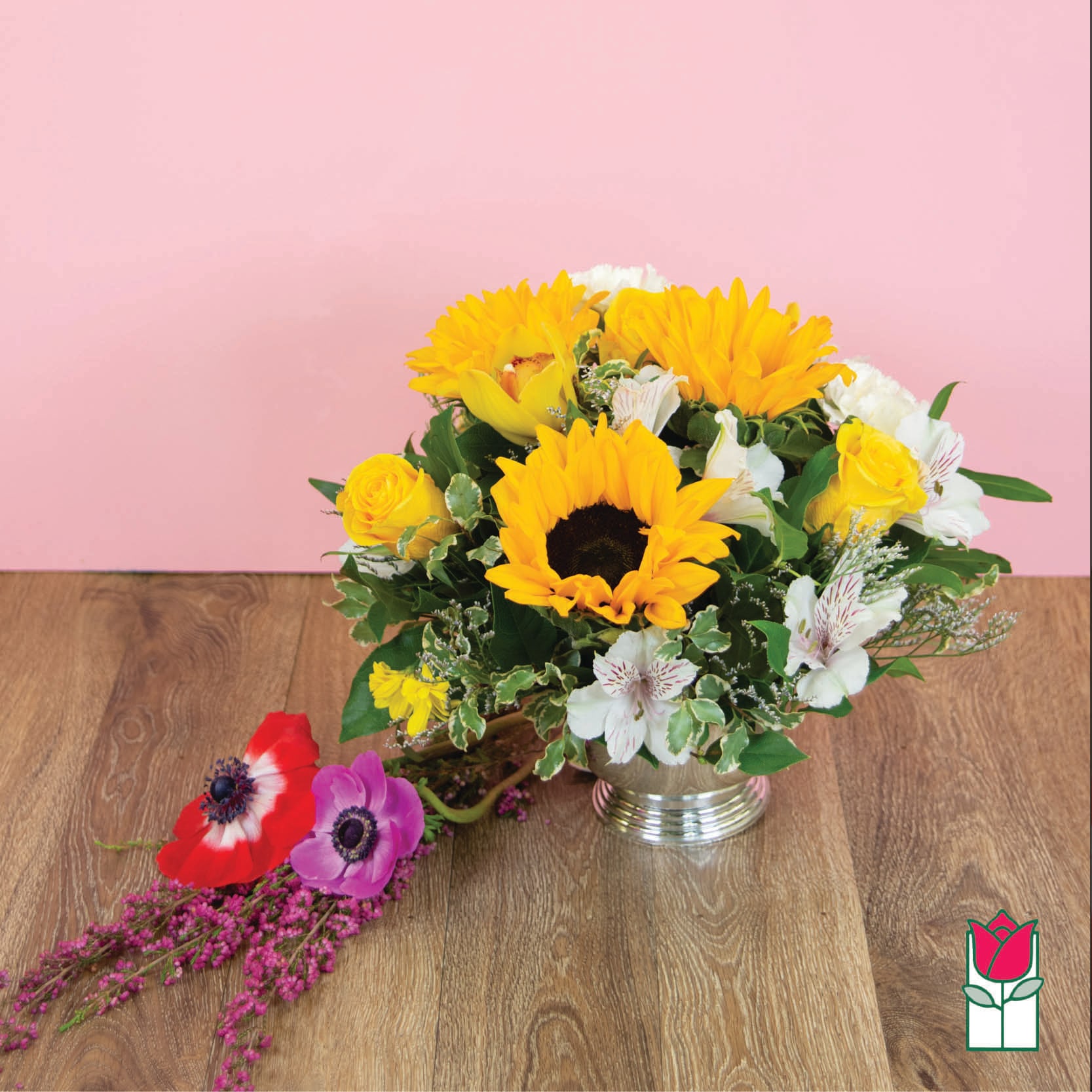 Yellow sunflowers and white flowers in a silver vase with loose blooms beside it.