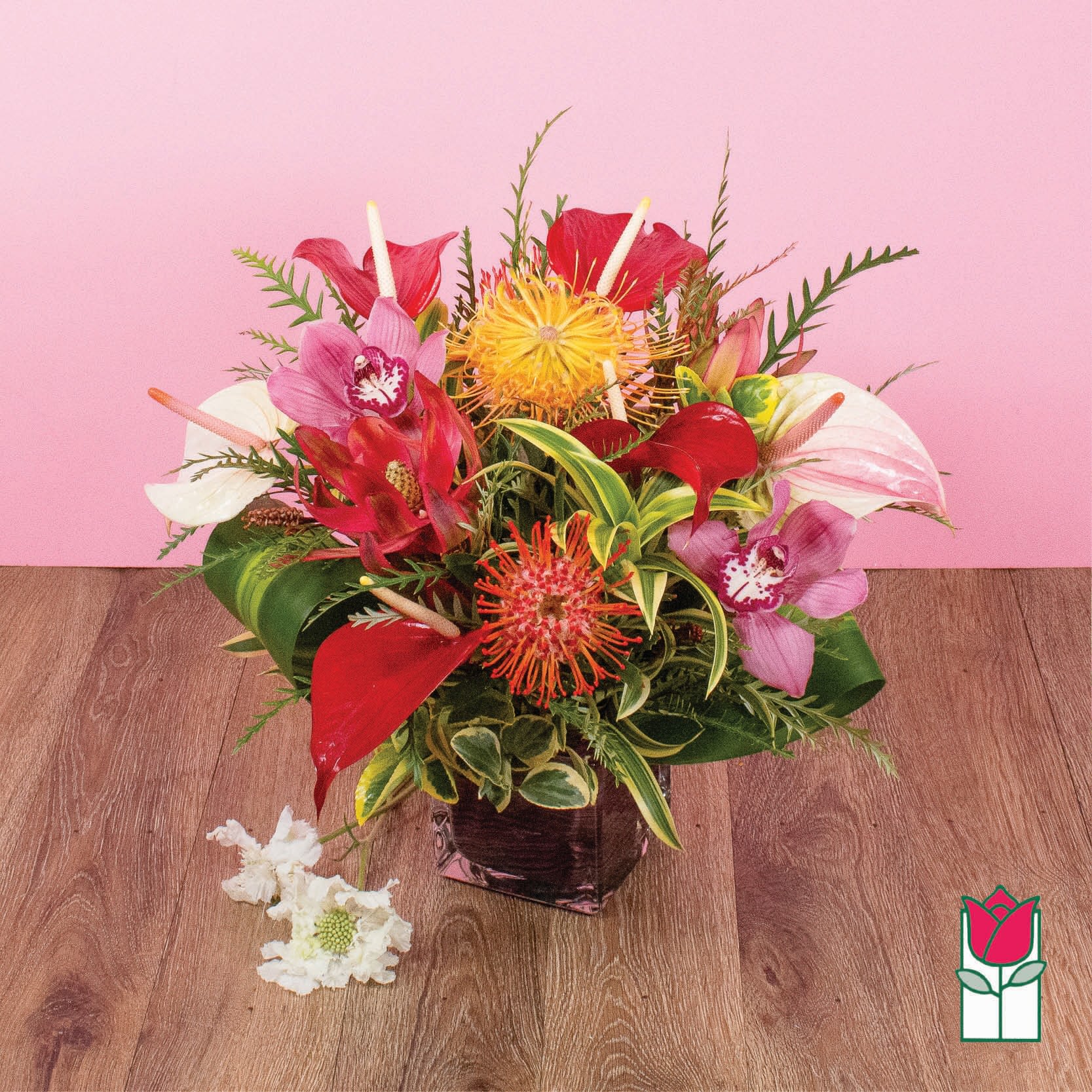 Tropical arrangement with red anthuriums, pink orchids, and yellow protea in a square glass vase.