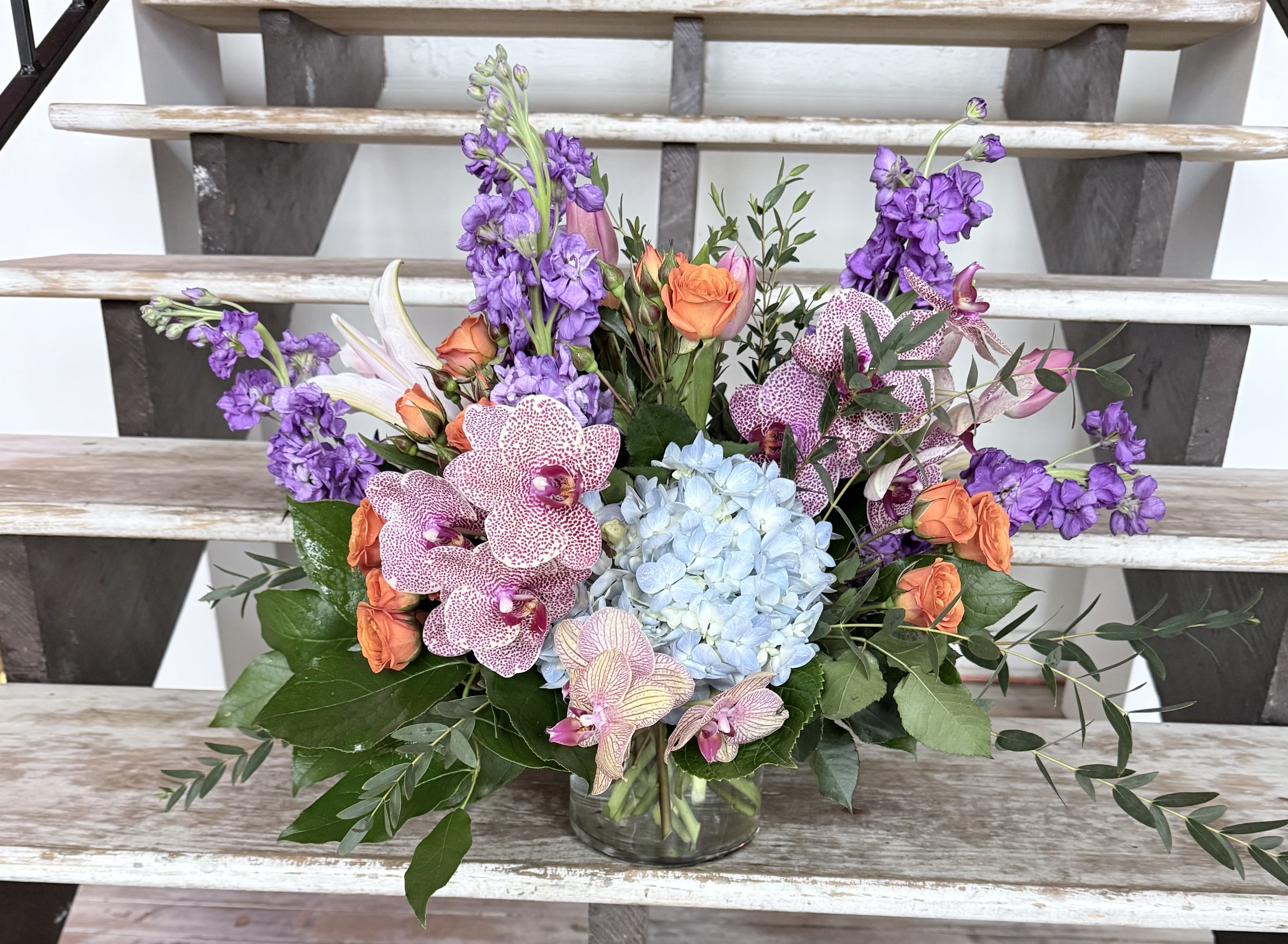 Mixed bouquet with purple blooms, orange roses, blue hydrangea, and orchids in a glass vase