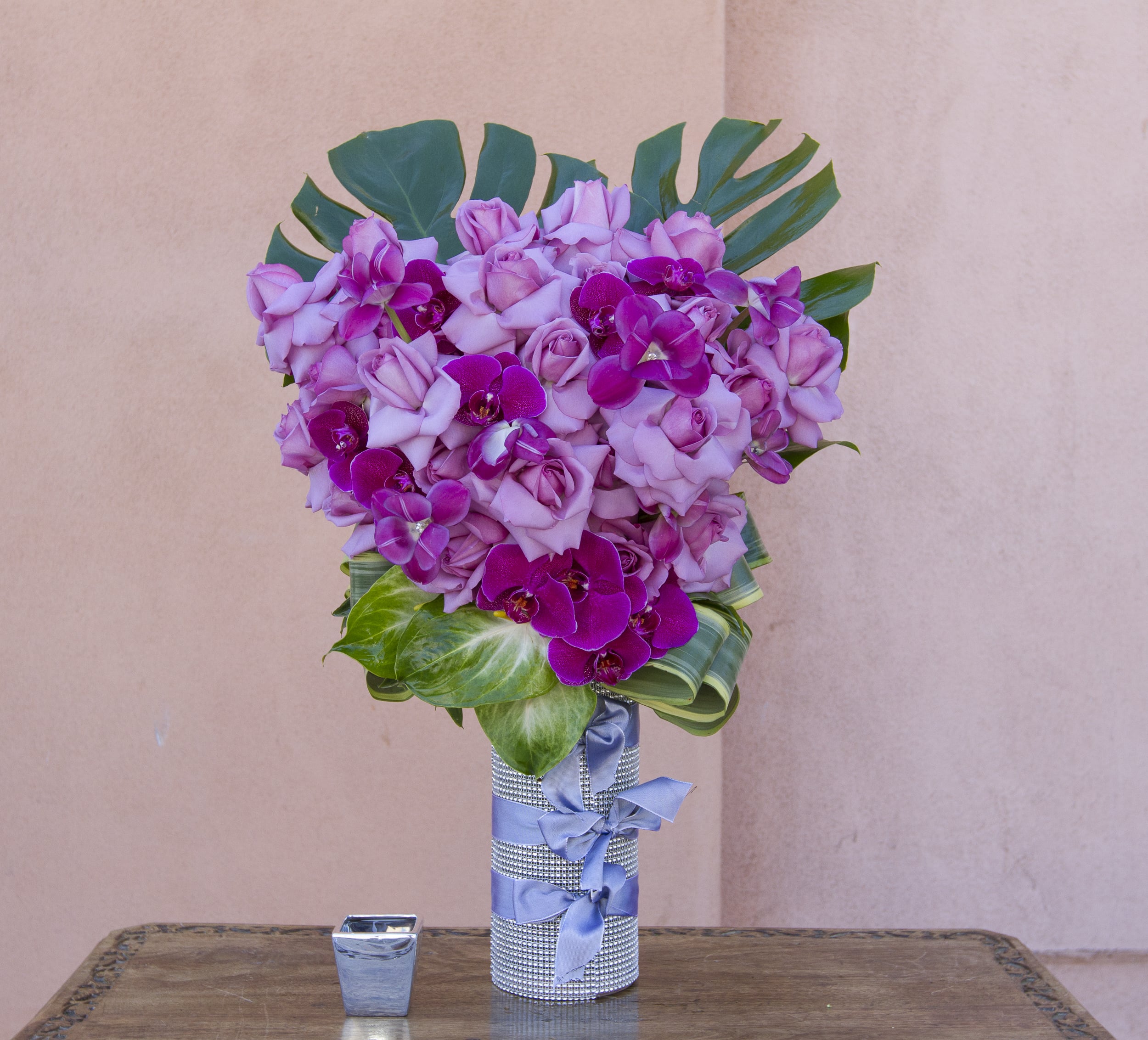 Pink roses and purple orchids arranged in a silver vase with a blue ribbon