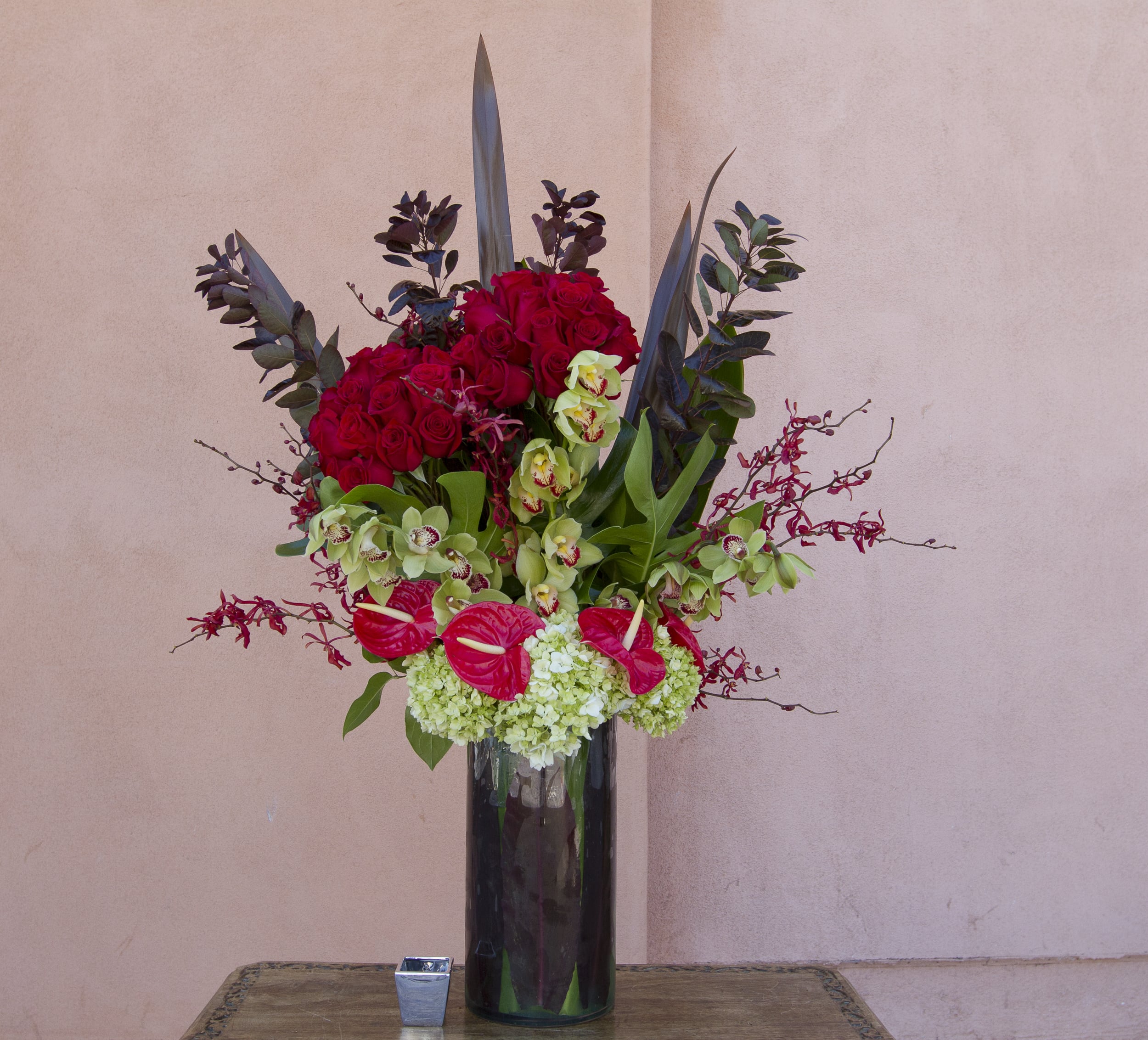 Tall arrangement of red roses, green orchids, and anthuriums in a dark vase