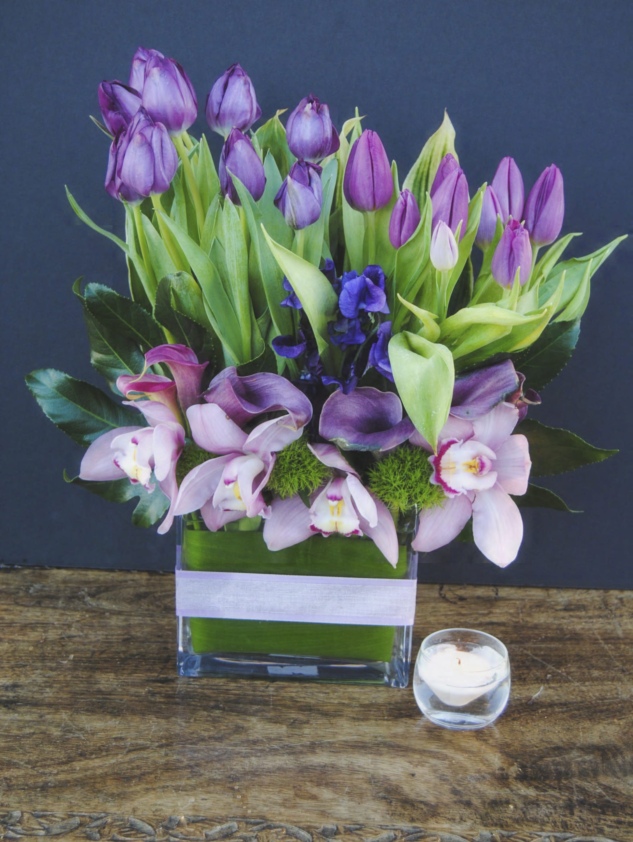 Purple tulips and pink orchids arranged in a glass vase with a small candle nearby