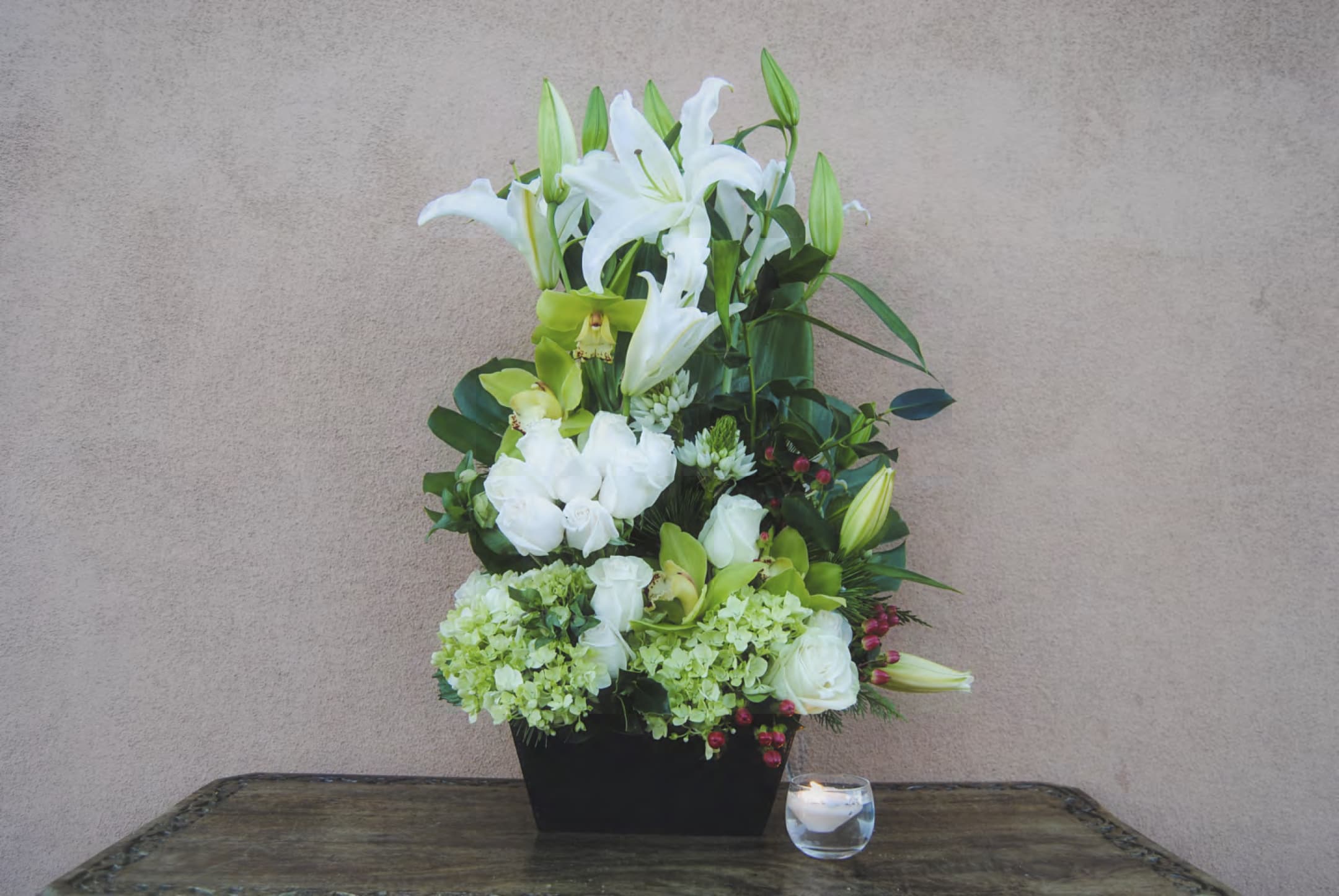 White lilies and green blooms in a black container beside a candle