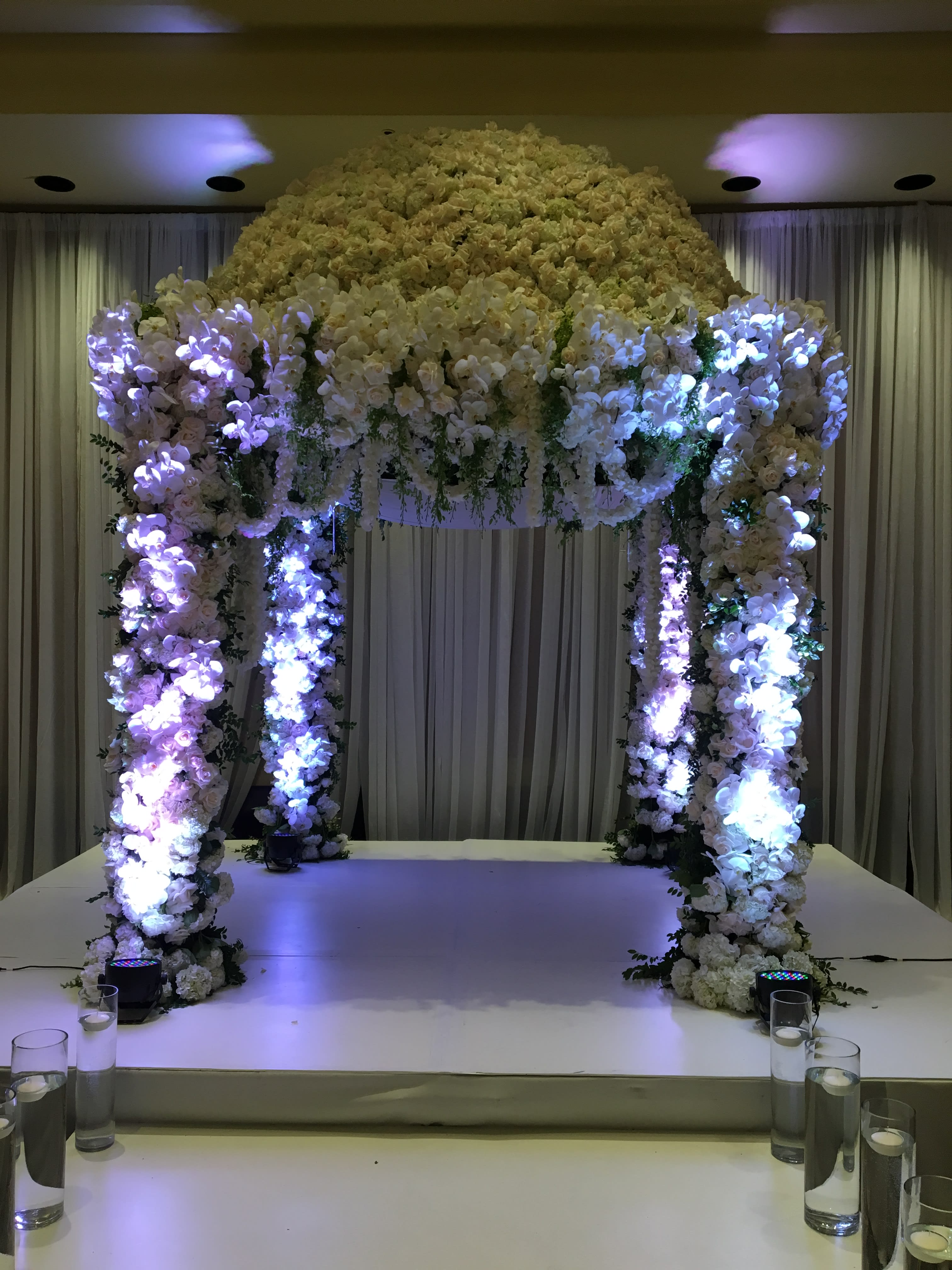 Large white floral wedding arch on a stage with purple lighting
