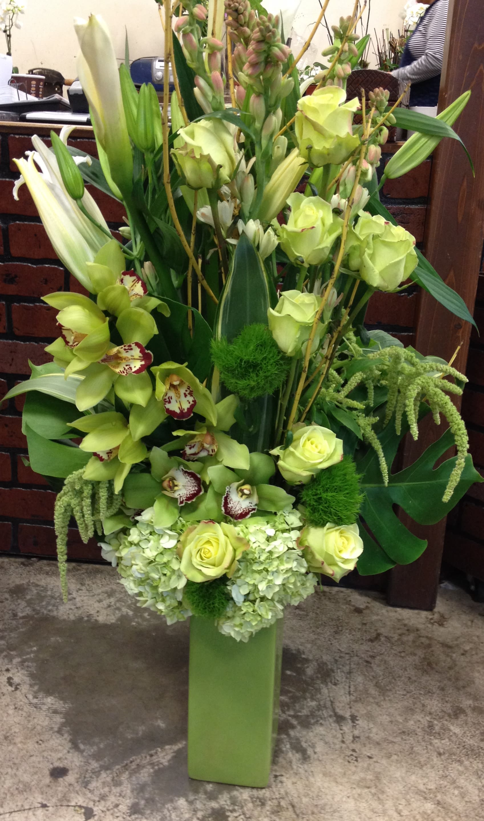 Tall green floral arrangement with orchids, roses, lilies, and hydrangeas in a rectangular vase