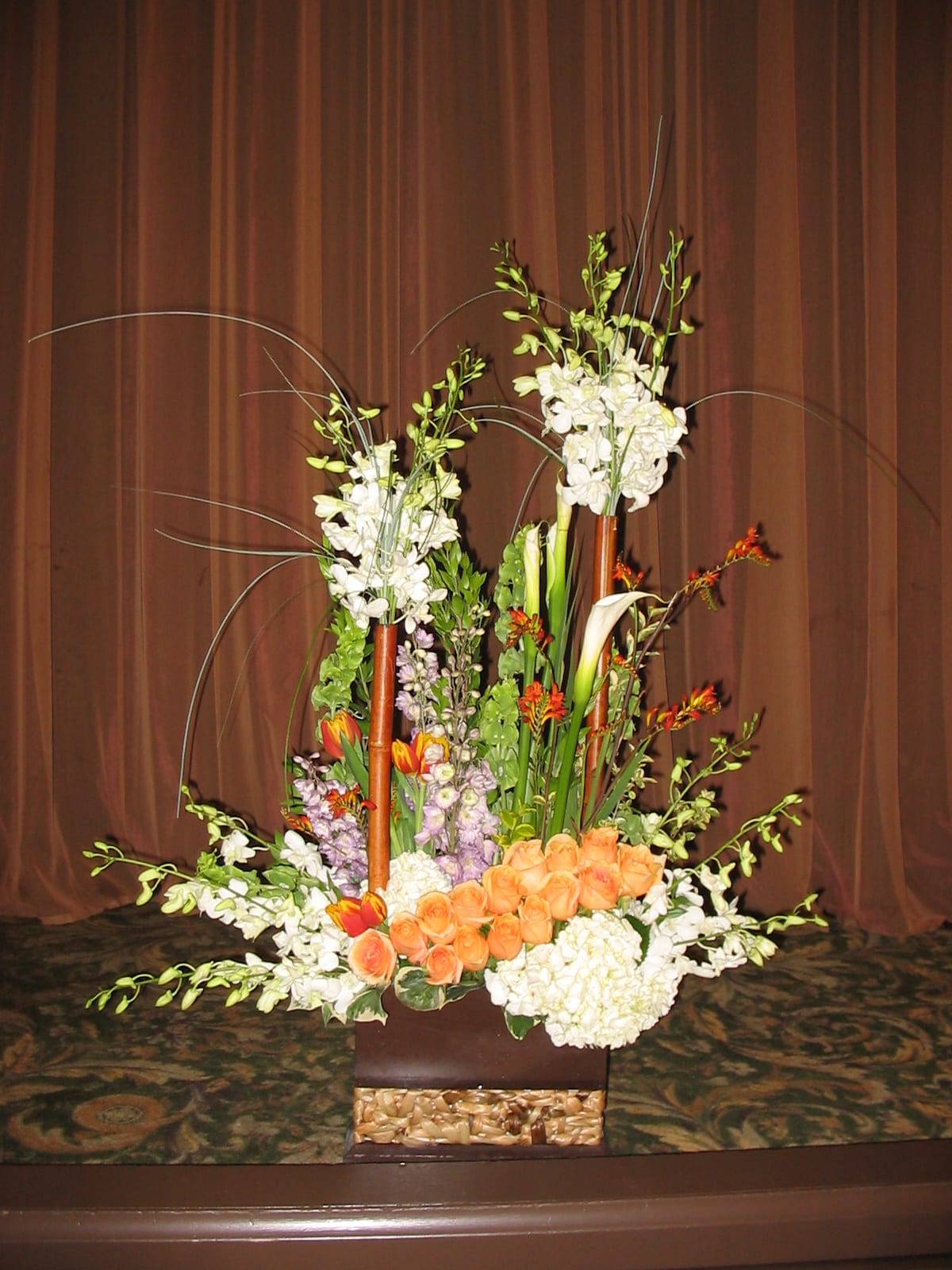 Tall floral arrangement with orange roses and white orchids in a basket vase