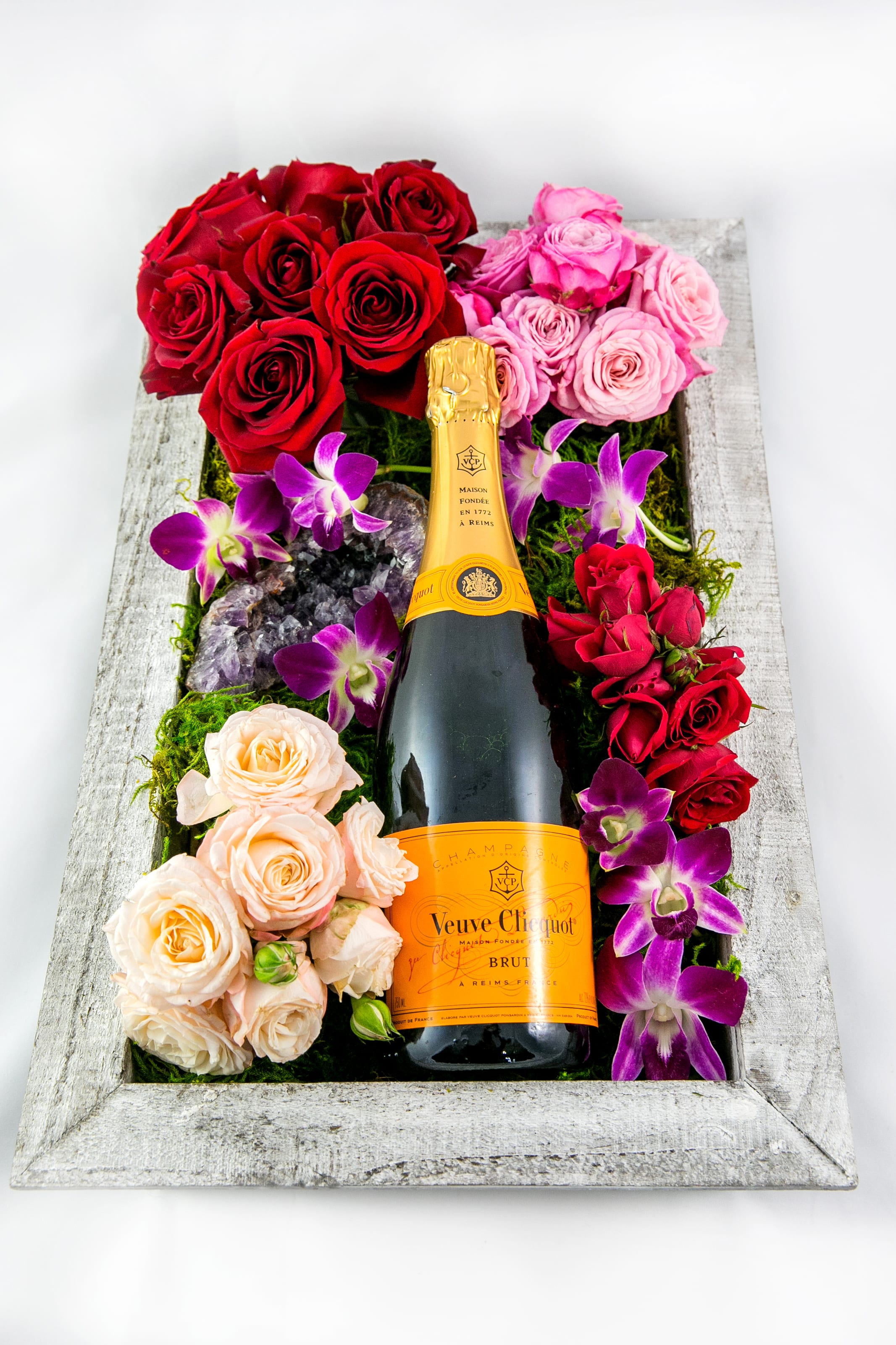 Champagne bottle surrounded by red, pink, and peach roses in a wooden box
