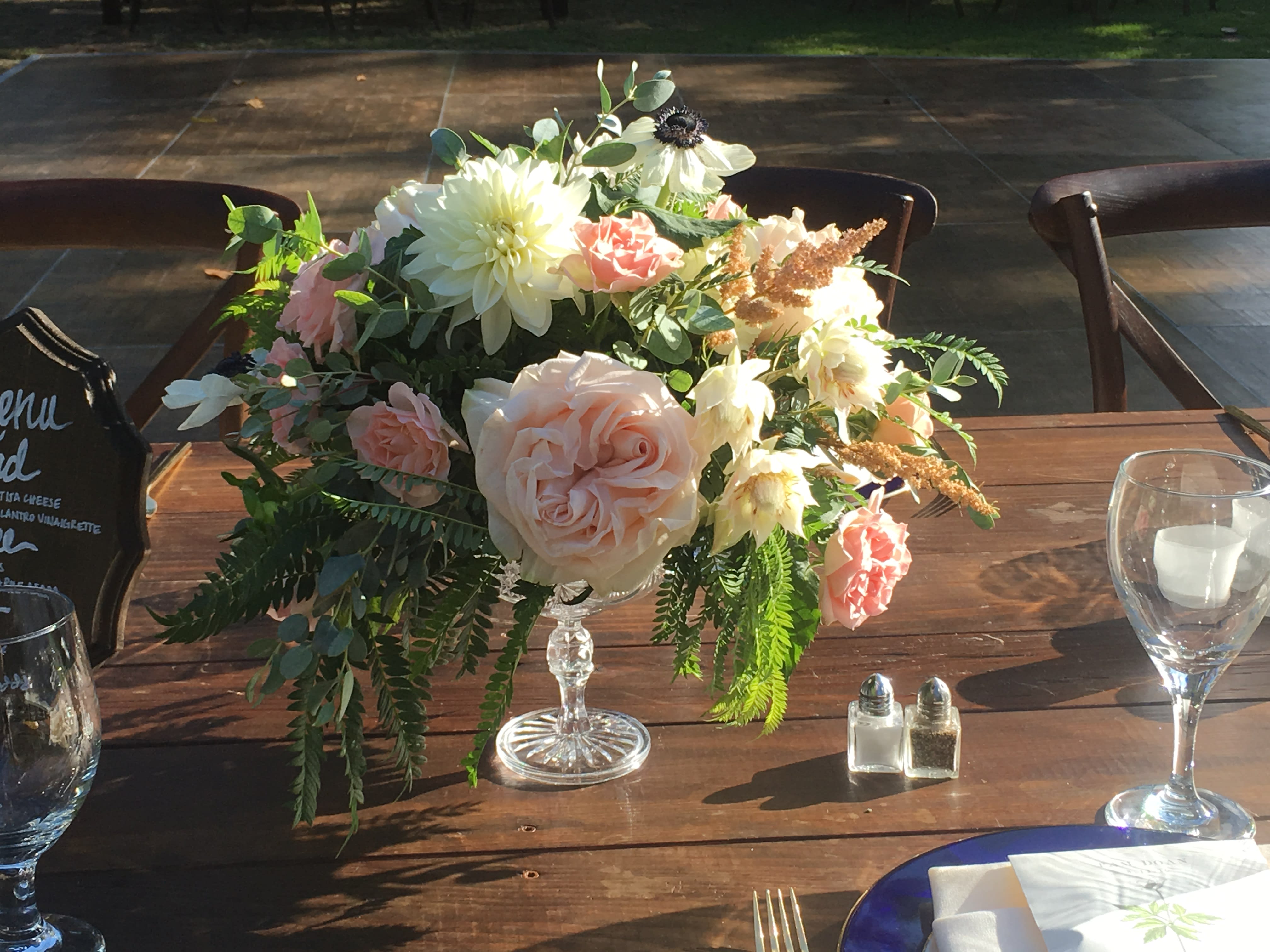 Pastel floral centerpiece in a clear pedestal vase on a wooden table