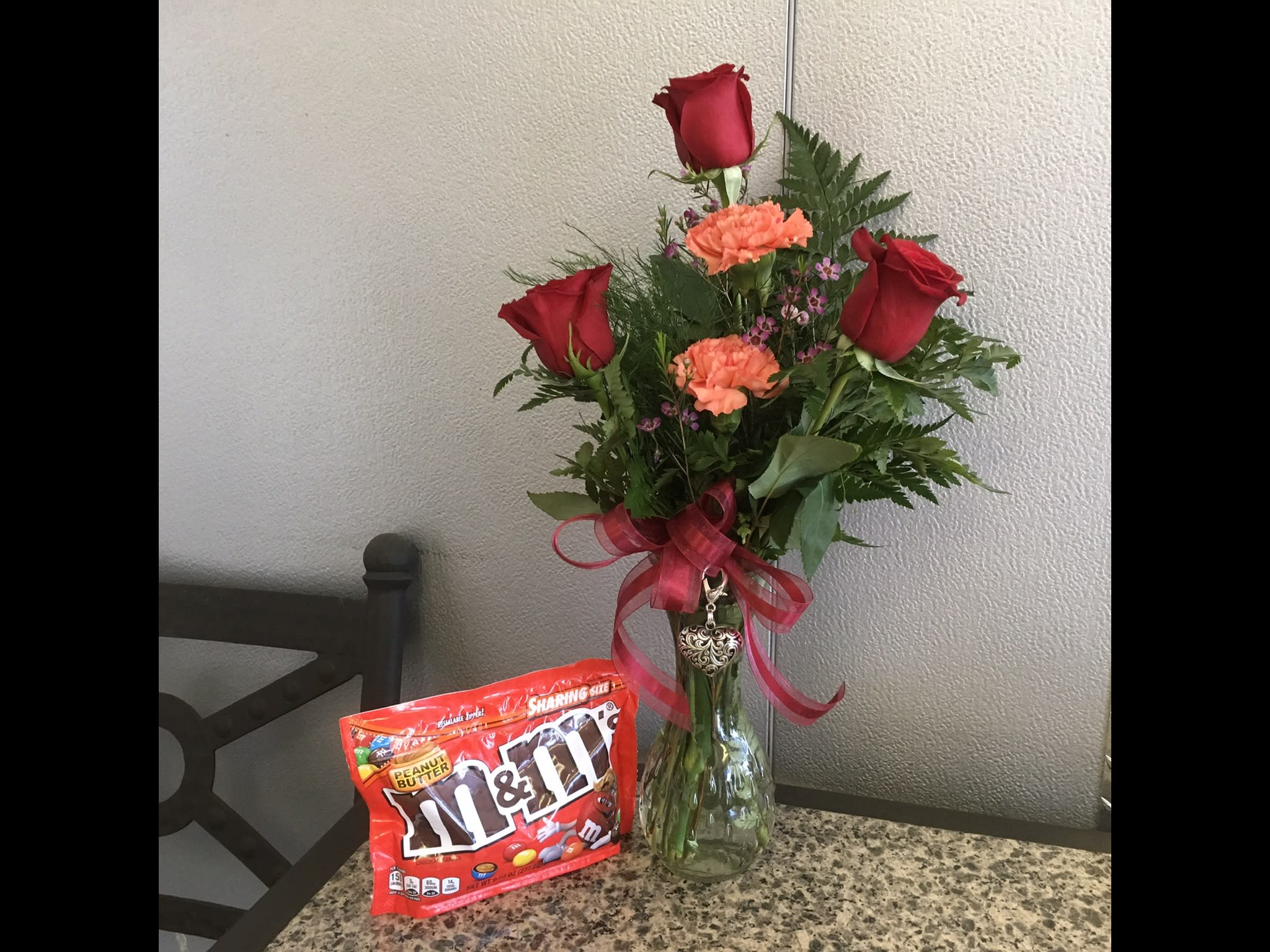 Red roses and orange carnations in a glass vase with a ribbon, beside a bag of M&amp;M's.