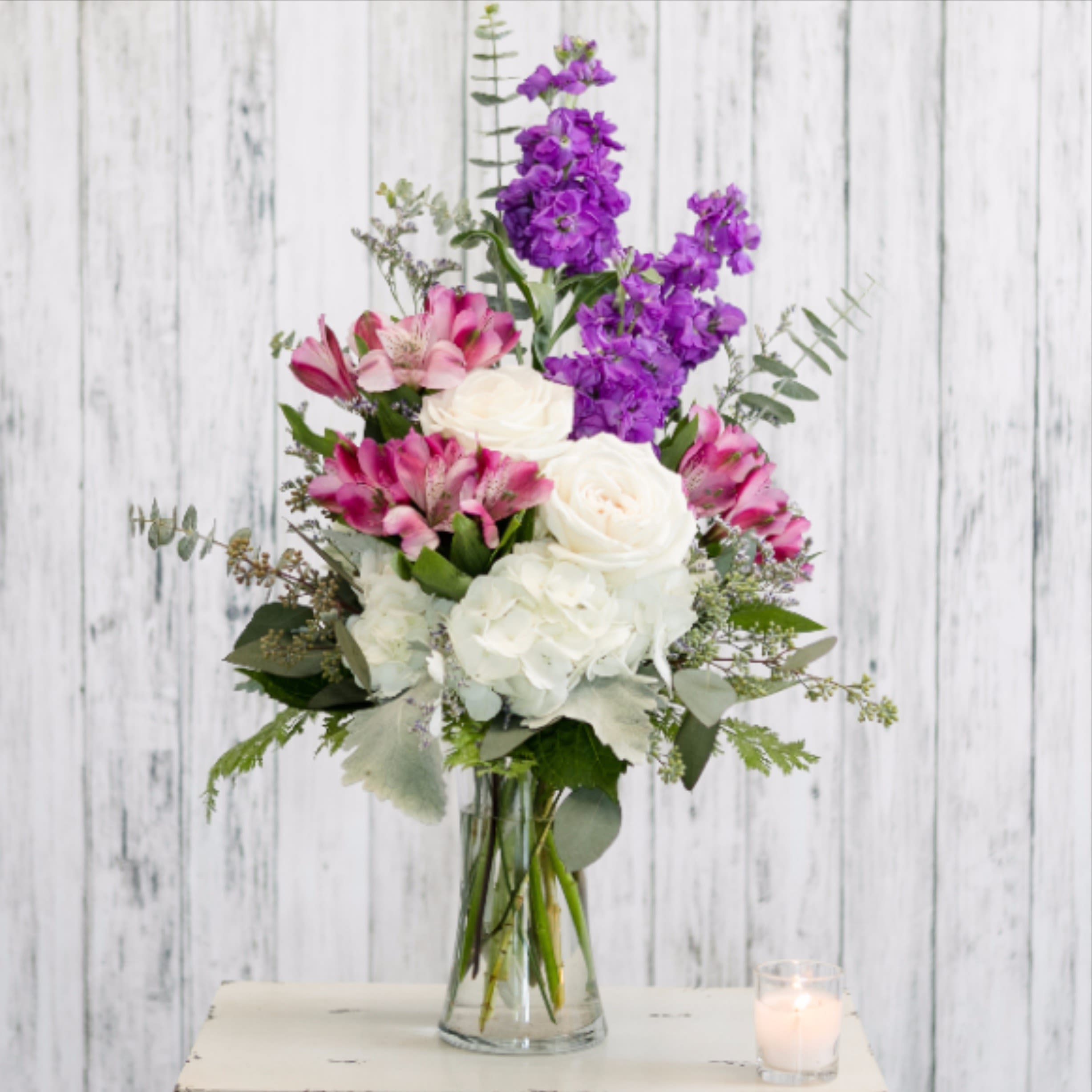 Tall mixed arrangement of white roses and hydrangeas with purple stock and pink alstroemeria in a glass vase