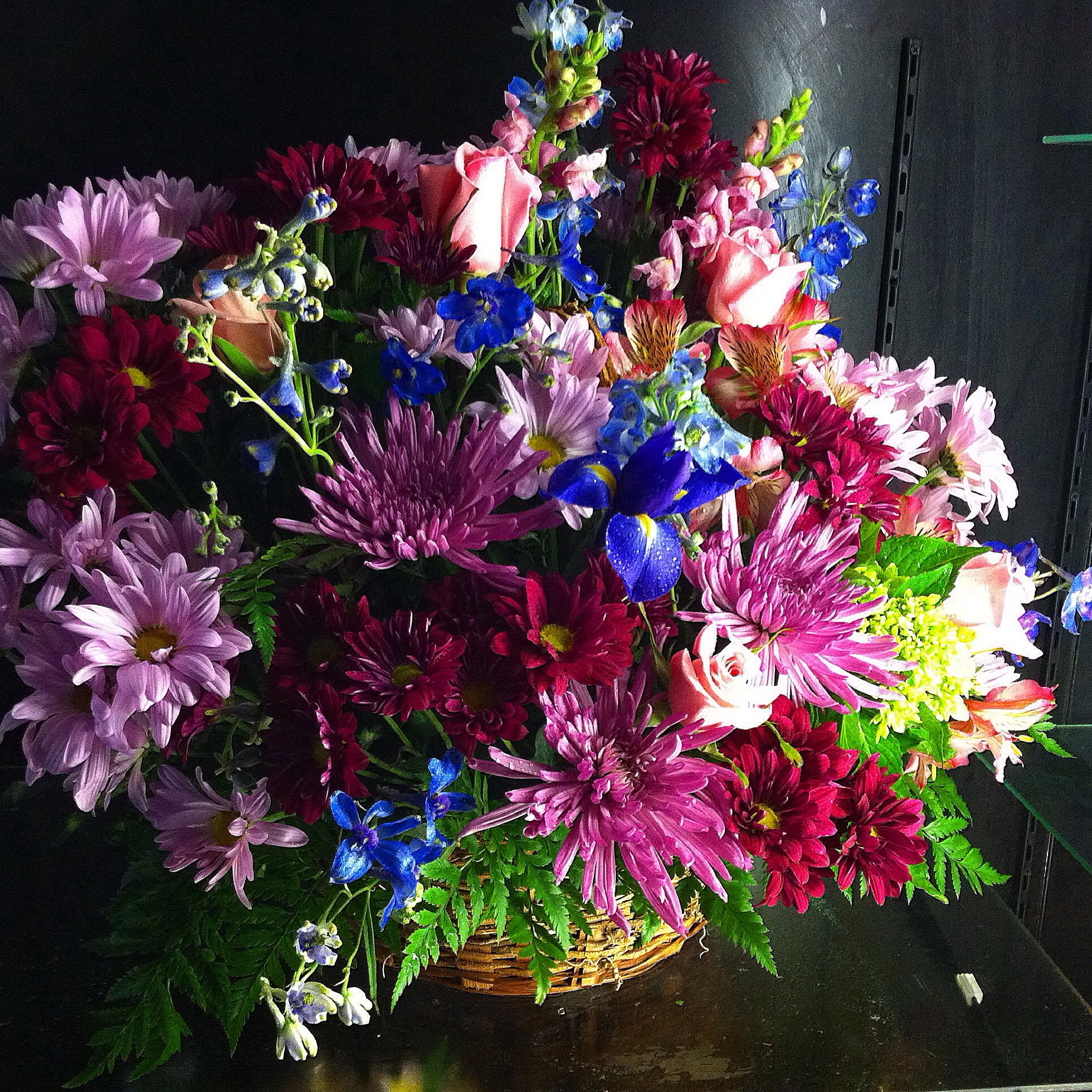 Mixed basket arrangement with pink roses, purple mums, and blue accent flowers in a wicker basket