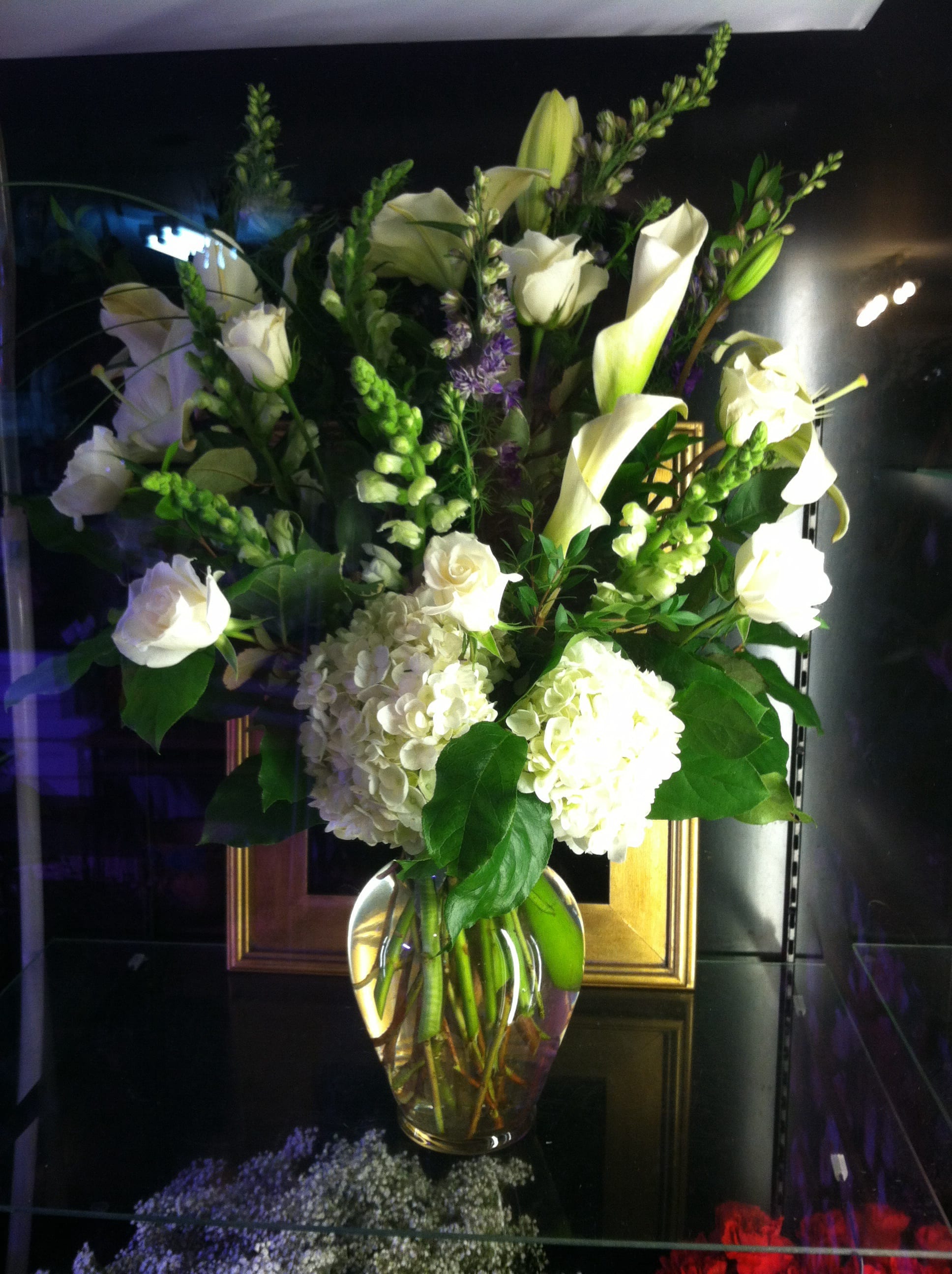 Tall arrangement of white roses, calla lilies, and hydrangeas in a clear glass vase with touches of purple.
