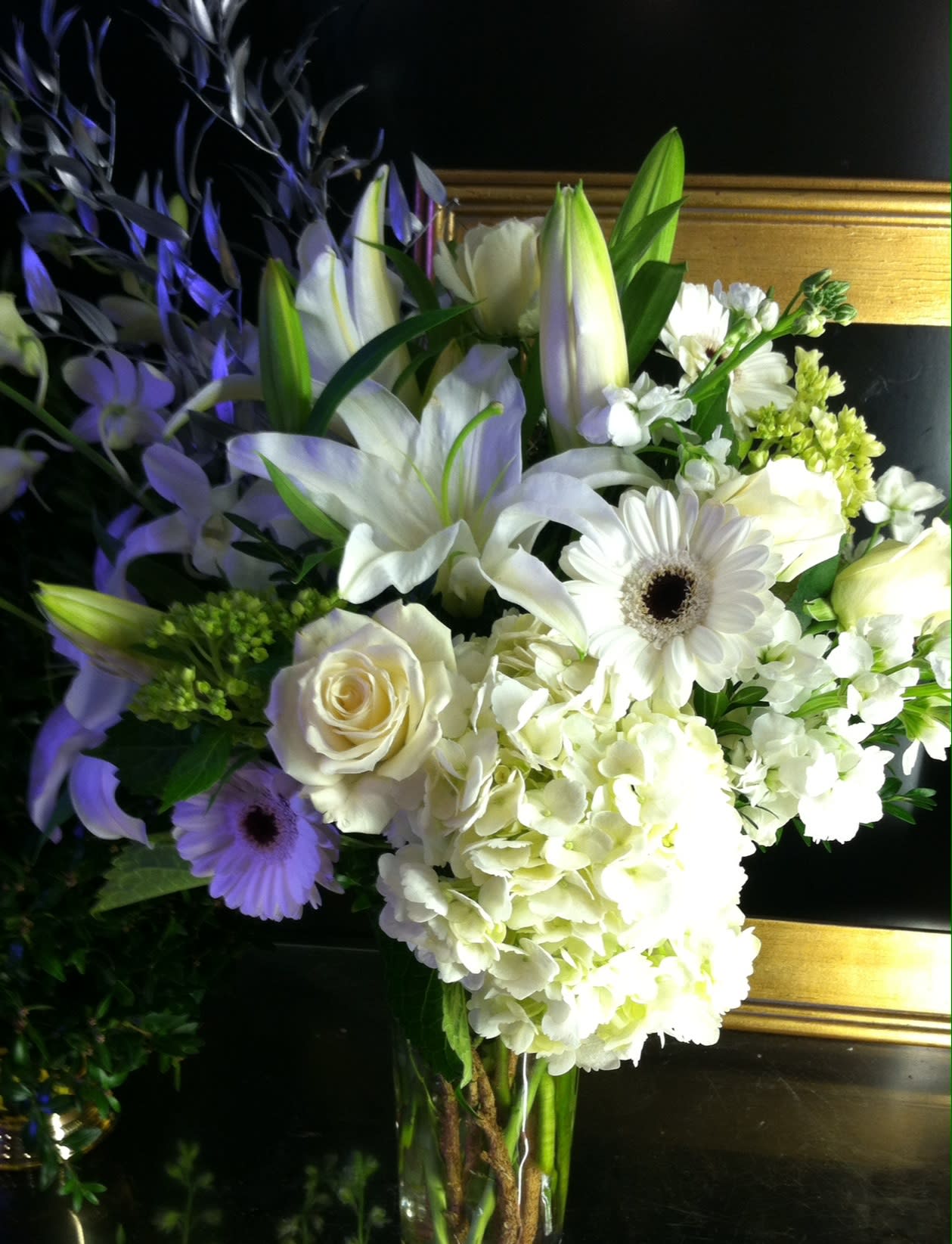 Tall white floral arrangement with lilies, roses, hydrangeas, and gerbera daisies in a clear glass vase