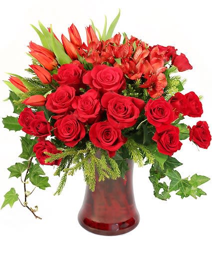 Arrangement of red roses, tulips, and alstroemeria in a red glass vase with trailing ivy.