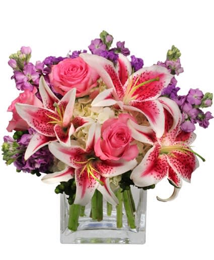 Compact arrangement of pink lilies, pink roses, and purple flowers in a clear glass cube vase