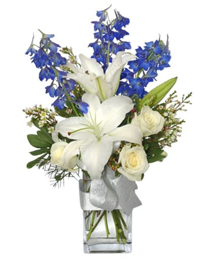 Tall arrangement of white lilies and roses with blue flowers in a clear vase with a silver ribbon.