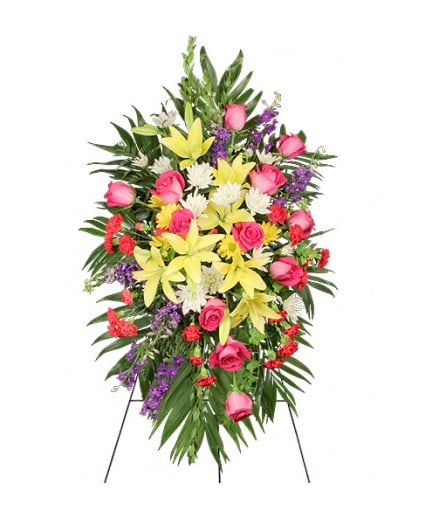 Standing spray of yellow lilies, pink roses, red carnations and purple flowers on lush green foliage.