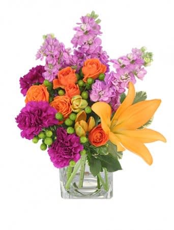 Bright arrangement of orange lilies and roses with purple flowers in a clear square vase