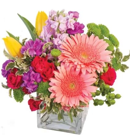 Colorful mixed bouquet with pink gerbera daisies, red roses, purple blooms, and yellow tulips in a glass cube vase