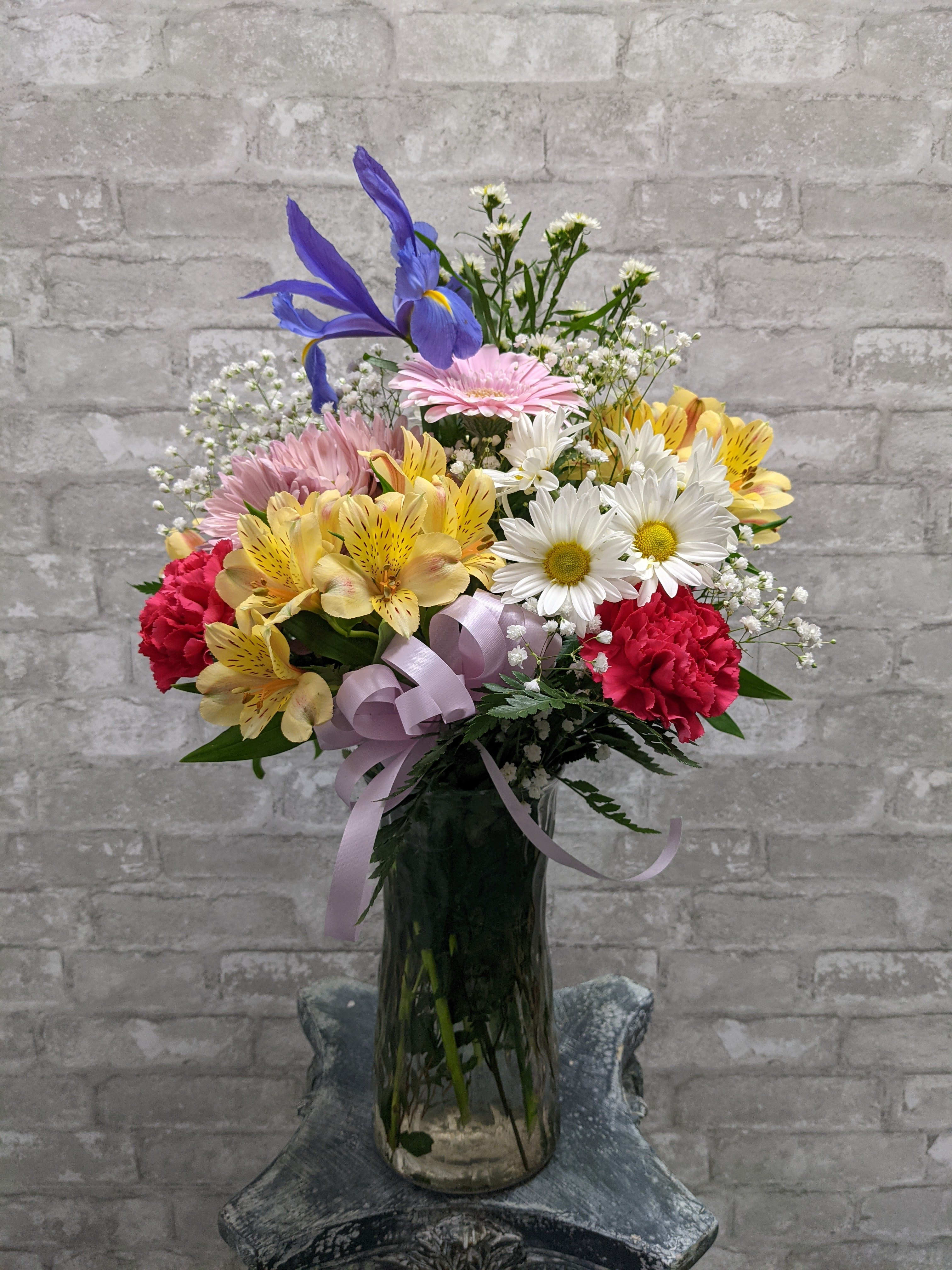 Mixed bouquet of colorful flowers in a tall glass vase with a lavender ribbon