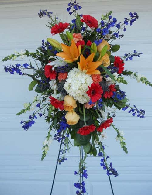 Tall mixed floral spray with orange lilies, red daisies, and blue flowers on an easel