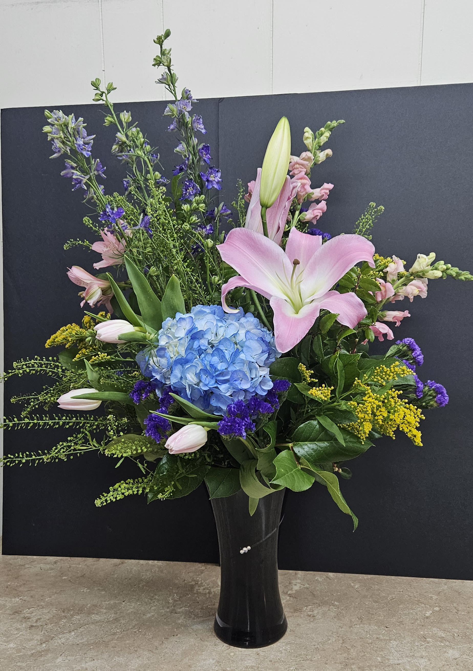 Tall mixed bouquet with pink lilies, blue hydrangea, and purple flowers in a black vase