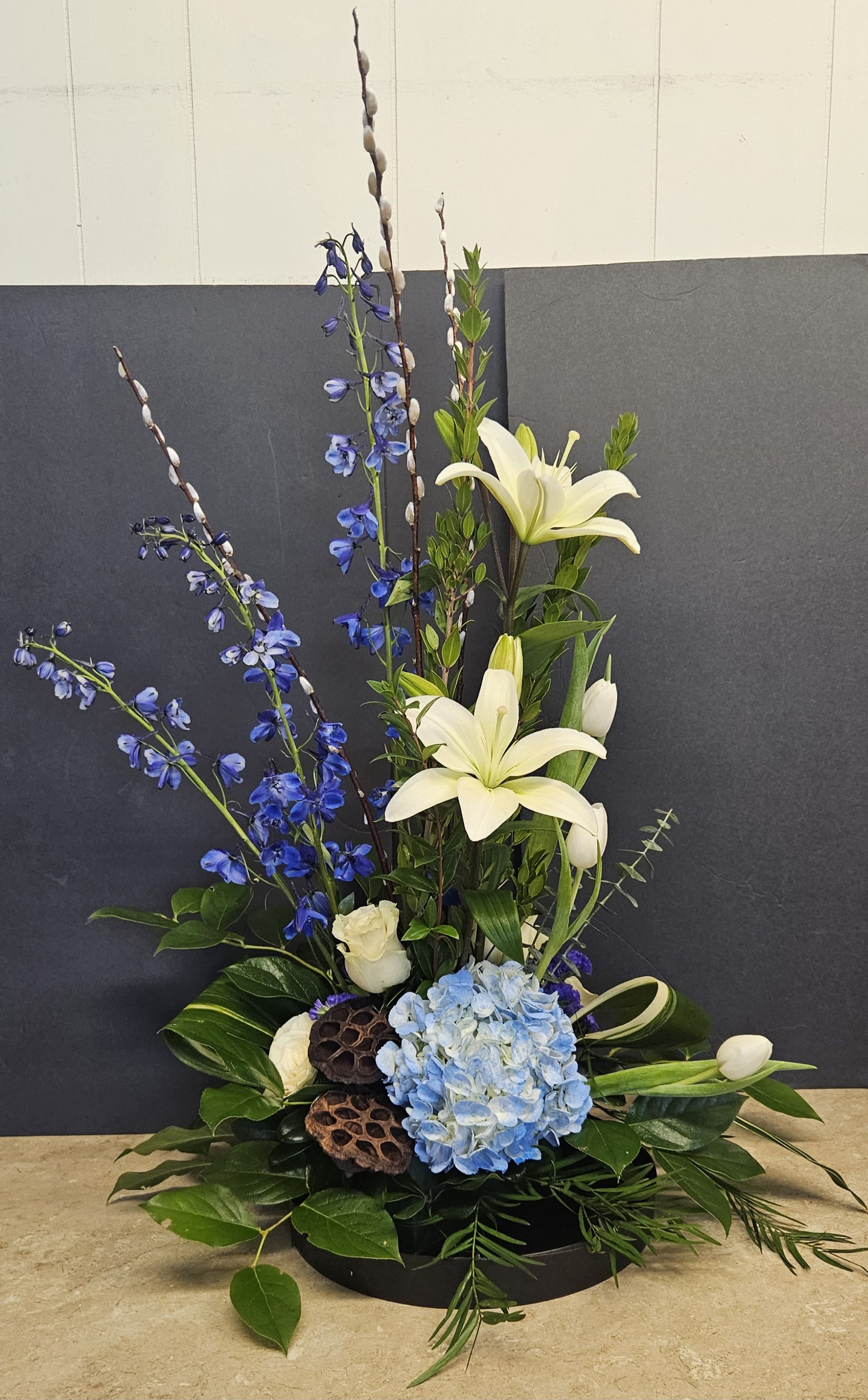 Tall floral arrangement with white lilies, blue flowers, and hydrangea in a low black base