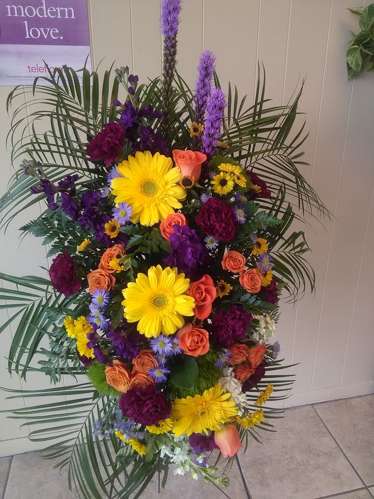 Tall standing floral spray with yellow daisies, orange roses, and purple blooms