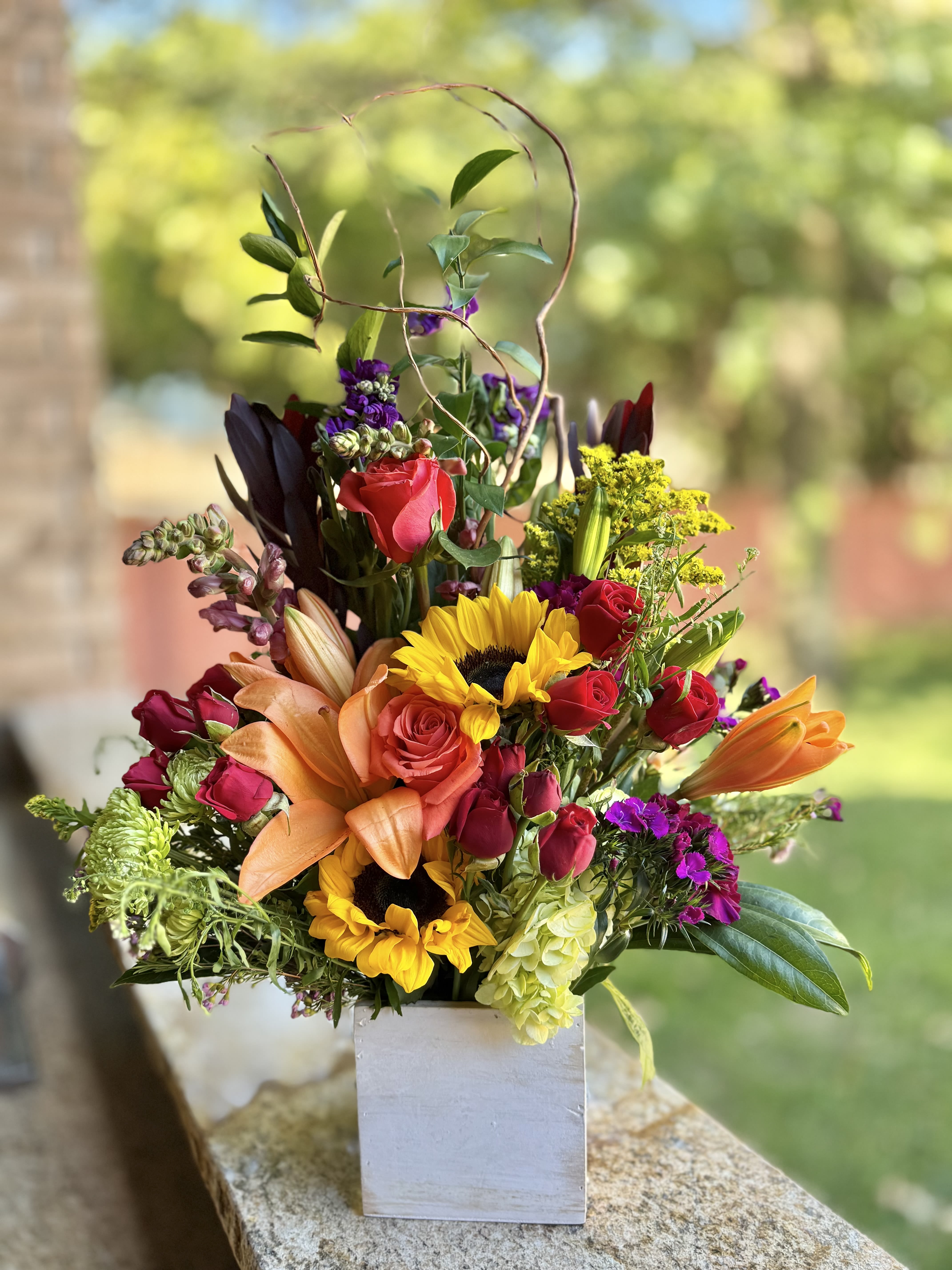 Mixed bouquet in a white square vase with roses, sunflowers, lilies, and purple blooms