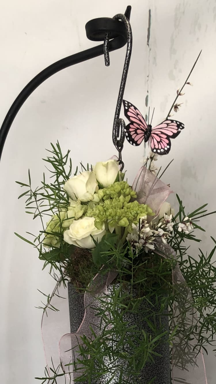 White roses and green blooms in a hanging metal basket with a pink butterfly decoration.