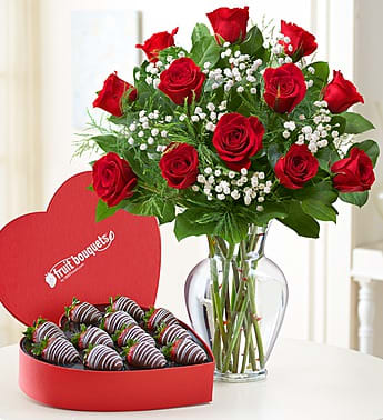 Red roses in a glass vase beside a heart-shaped box of chocolate-covered strawberries.
