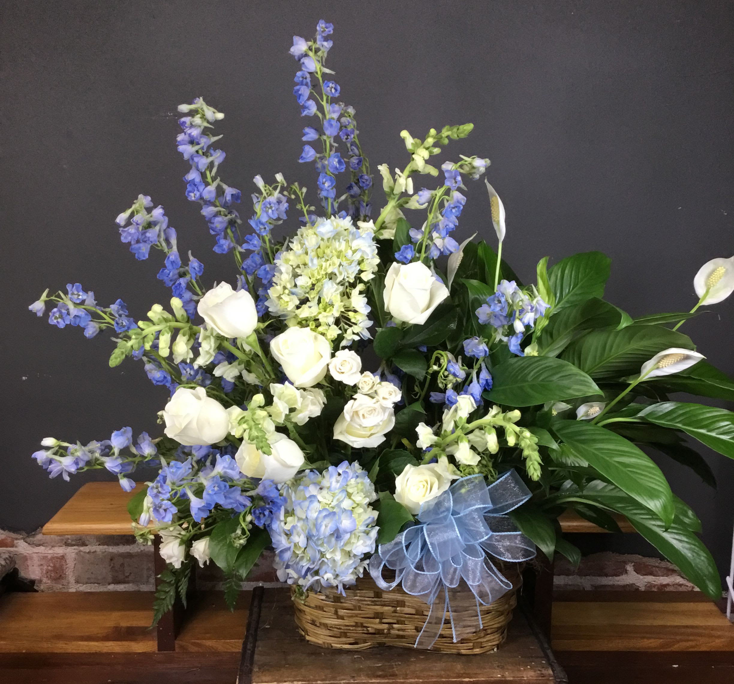 Peacful and Blue - Cool, airy whites and pale blues set a quietly elegant tone in this flower arrangement-the perfect sympathy arrangement, wedding centerpiece, or thank-you gift to comfort and uplift. White roses and pale blue hydrangeas create a soft, romantic base while tall delphinium spikes and white snapdragons add vertical drama; the overall impression is serene and thoughtfully formal, designed to make the recipient feel genuinely seen and comforted.  Arranged in a low wicker basket with a sheer blue ribbon, the composition pairs sculptural white calla lilies with glossy broad foliage that fills and balances the right side. Expect gentle textures from the hydrangea heads, the velvety smoothness of roses, and the architectural lines of delphinium and snapdragons-an arrangement that reads refined without feeling fussy. The basket presentation makes it ready to give, whether you're honoring a solemn moment, celebrating a quiet wedding, or saying thank you with understated grace.