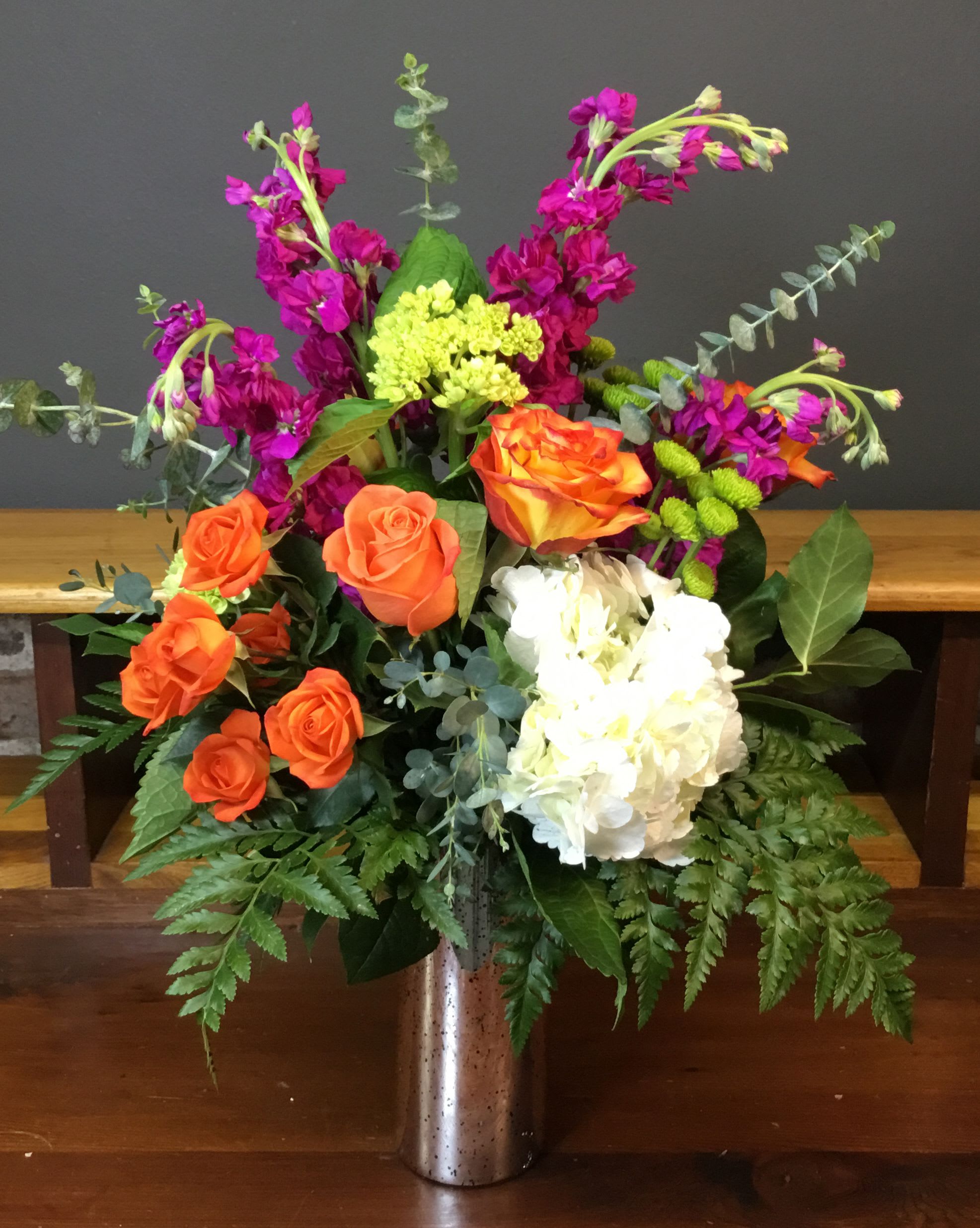 Show Stopper - A bold burst of color-this mixed-color flower arrangement pairs warm orange roses with a large white hydrangea cluster and vivid magenta spiky blooms to create an energetic, garden-style bouquet perfect for a birthday bouquet, congratulations gift, or cheerful thank-you. The upright, full composition and lively silhouette make the recipient feel truly celebrated the moment they see it.  The design includes roses, hydrangeas and mixed seasonal blooms set among airy eucalyptus and fern foliage, arranged in a modern metal vase for a polished, gift-ready presentation. Tall stems reach outward for movement and texture while the contrasting orange, white and bright pink palette keeps the look fresh and joyful.  This is the kind of arrangement that announces happy news and brightens a room-ideal for friends, coworkers, or anyone who loves vivid color. Vase is included; no special care beyond fresh water and a cool spot will keep blooms looking great through the week.