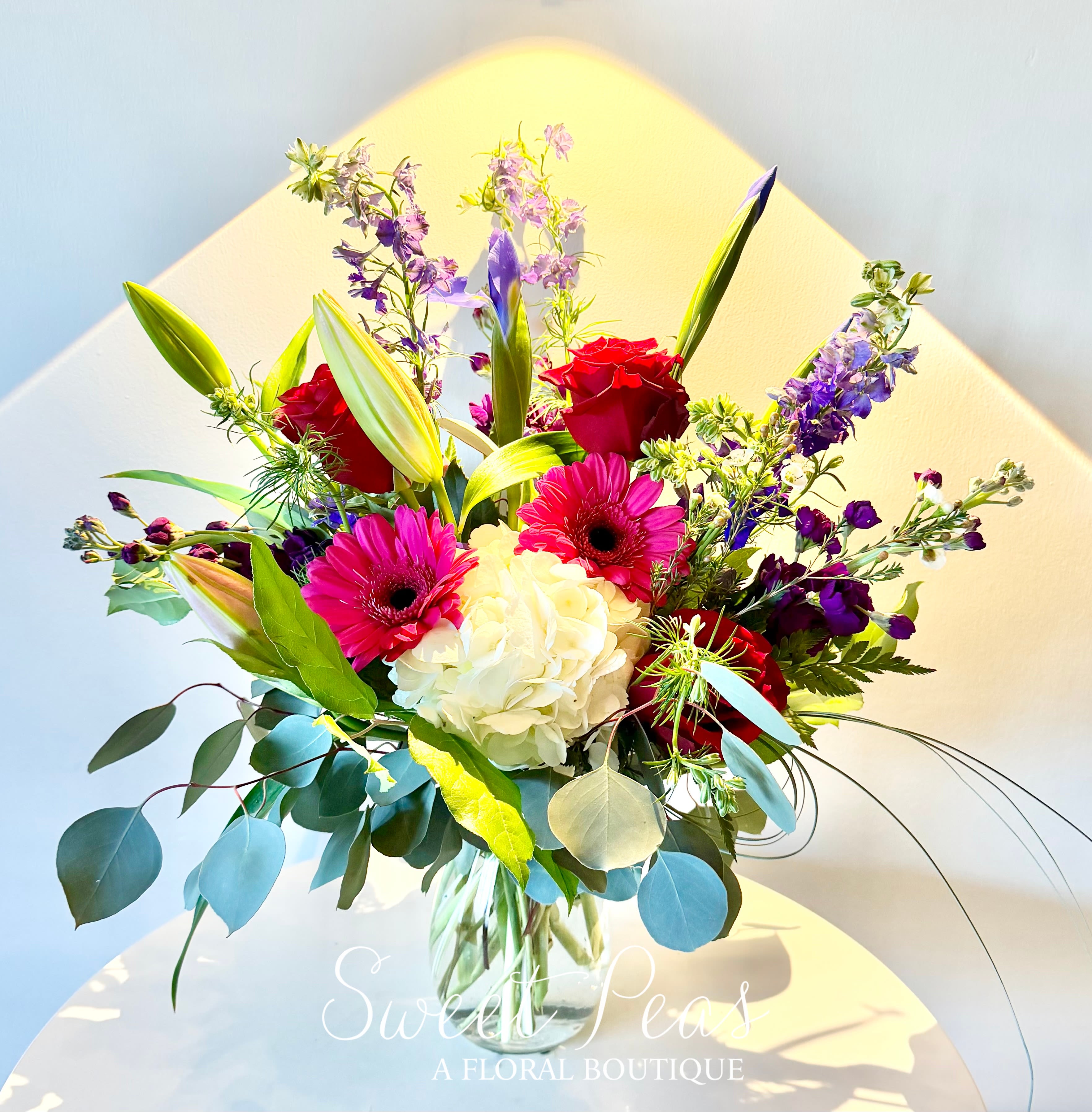 Bouquet of red roses, pink gerbera daisies, white hydrangea, and purple blooms in a glass vase