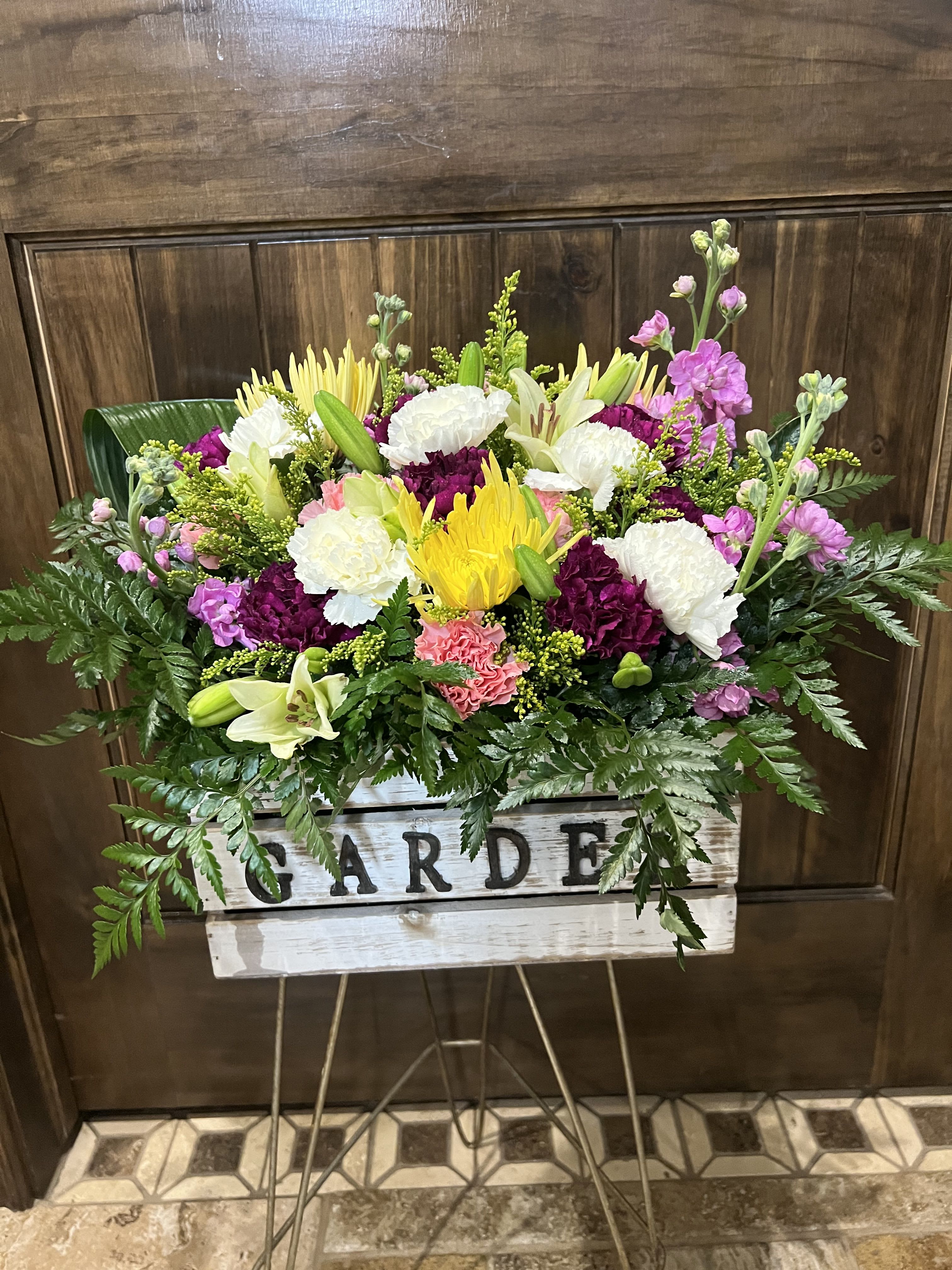 Spring Garden  - A cheerful, garden-inspired flower arrangement that reads like a slice of spring - perfect as a Mother's Day bouquet, birthday surprise, or just-because gift. This rectangular whitewashed wooden box labeled "GARDEN" holds a wide, full display that brings together roses, lilies, carnations, chrysanthemums and fragrant stock in a lively mix of purple, pink, white, yellow and fresh greens. The overall effect feels bright and hopeful, an ideal choice for Easter, get well wishes, housewarming, or to celebrate an anniversary with a touch of informal romance.  Clusters of deep purple and crisp white blooms gather at the center, highlighted by soft pink blossoms and sunny yellow spider-style flowers that add texture and movement. Abundant fern fronds frame the arrangement and spill over the edges of the box, creating a loose, garden-fresh silhouette that sits on a metal stand or tabletop. Sized to make an inviting statement without overwhelming a space, this mixed-color design sends a message of warmth and care - thoughtful for thanking a friend, lifting a coworker's spirits, saying "I'm sorry," or marking seasonal moments from Spring through Summer. Simple care: trim stems and refresh water as needed to keep the display looking freshly arranged.
