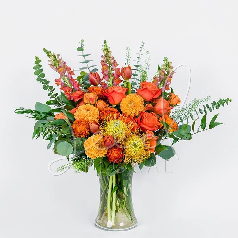 Tall arrangement of orange roses, marigolds, tulips and protea in a clear glass vase
