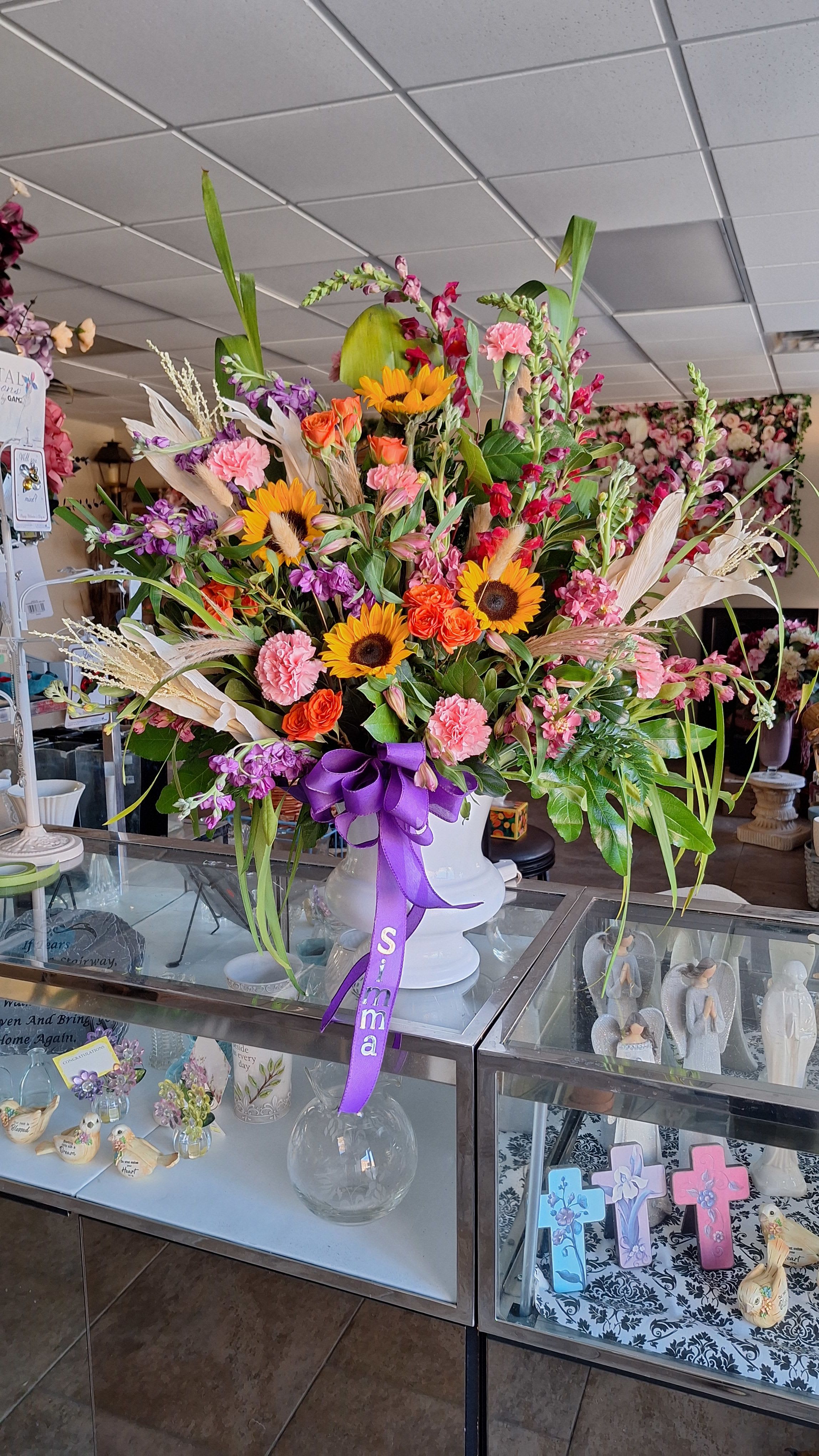Sun and Wheat Urn - Bright sunflowers and tall snapdragons give this funeral urn arrangement a striking vertical silhouette that celebrates a life with warmth and color. As a sympathy or funeral piece it brings sunlit yellow and orange cheer balanced by pinks and purples, and it also works beautifully as a wedding centerpiece or a bold gift to mark birthdays and congratulations-the person you're honoring will feel genuinely seen by its lively, uplifting presence.  This large mixed arrangement arrives in a white ceramic vase tied with a wide purple ribbon, combining sunflowers, roses, spray roses, alstroemeria, carnations, chrysanthemums, stock, snapdragons and seasonal wheat for rich texture and movement. The warm palette of yellow and orange is accented with pink and purple blooms to create depth; the tall spikes and fuller heads form a layered, abundant composition that reads as both celebratory and respectfully elegant.  Sized for a prominent display, the Sun and Wheat Urn fills a mantel, entry table, or reception setting with bold color and seasonal interest. Designed for easy care, flowers are arranged in floral foam inside the vase for stability-refresh water if possible and keep away from direct heat to prolong the life of the blooms.