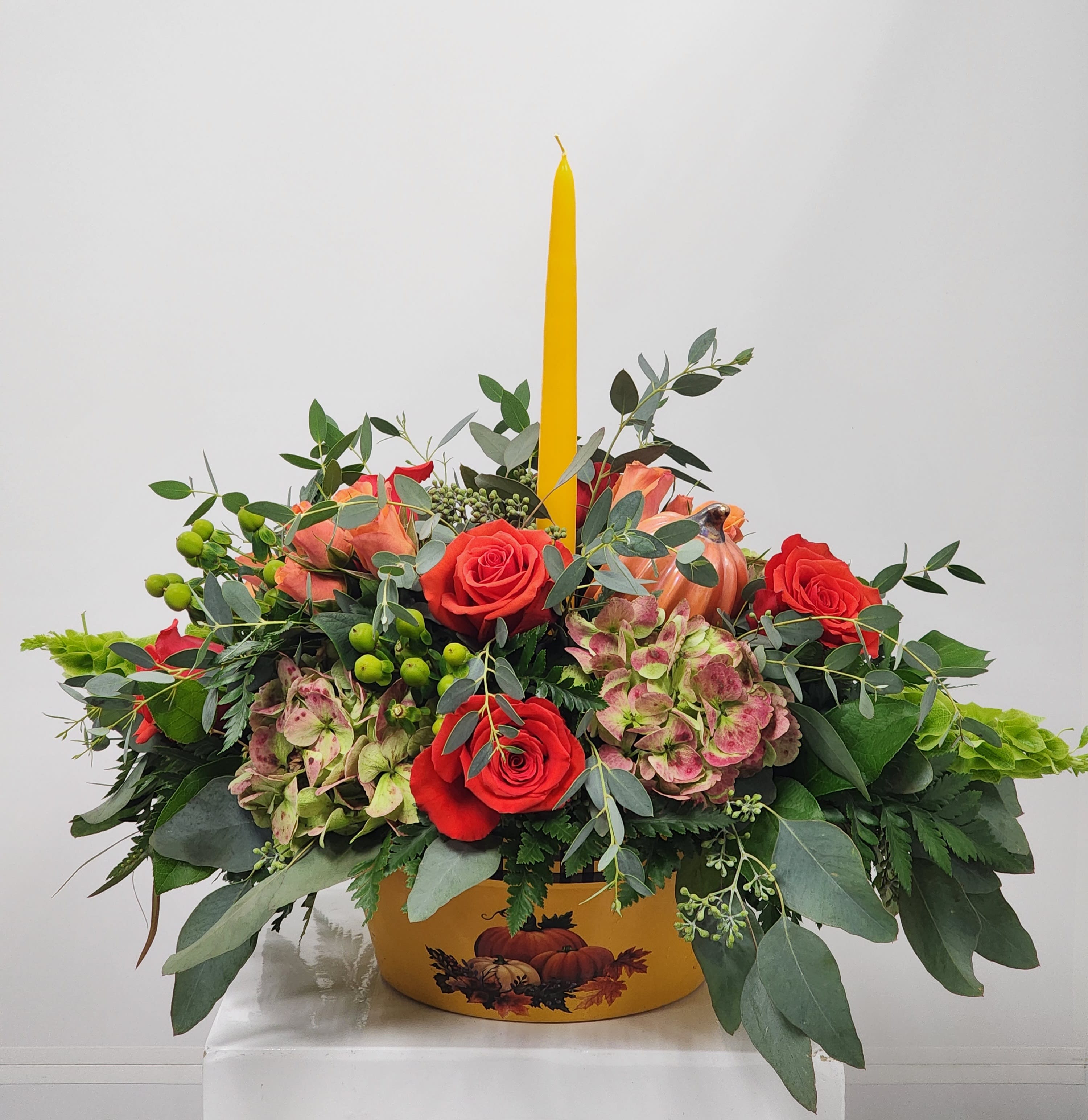 Amber Blooms - A rounded autumn centerpiece in an adorable yellow and plaid ceramic dish (candle included). Arranged with roses, spray roses, bells of Ireland, hypericum, and hydrangea.  Approximately 9.5" tall and 14" wide (without candle height added).