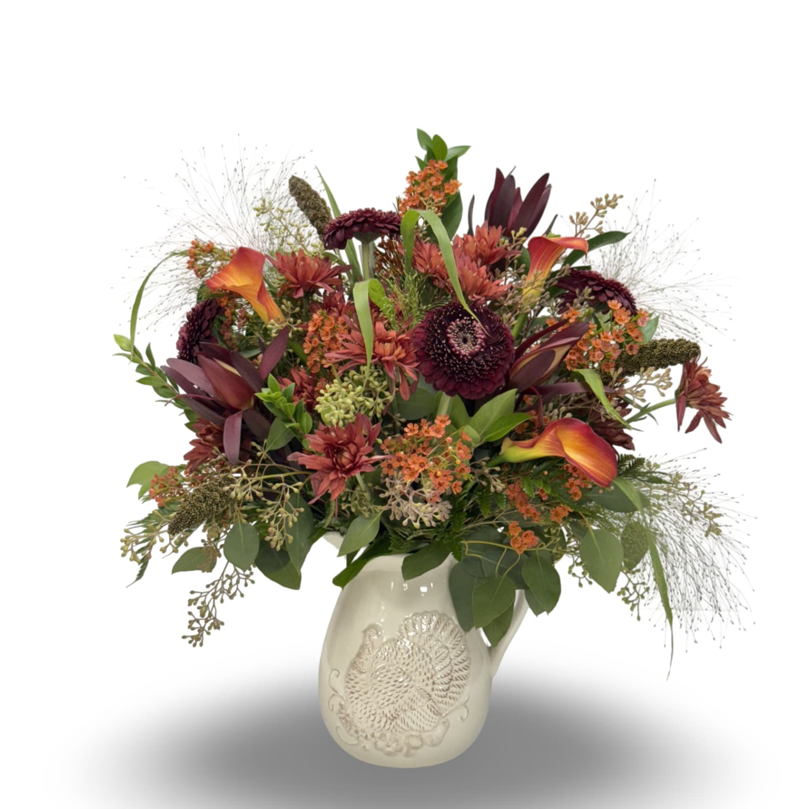 Country Corner - Bring the warmth of the harvest season into your home with our Country Corner arrangement. Designed in a charming ceramic pitcher embossed with a rustic turkey motif, this piece overflows with rich autumn tones. Featuring deep burgundy blooms, vibrant orange calla lilies, textured greens, and wisps of airy foliage, it captures the cozy elegance of fall. Perfect as a centerpiece or a heartfelt gift, this arrangement blends seasonal charm with timeless style—ready to be enjoyed long after the flowers fade.