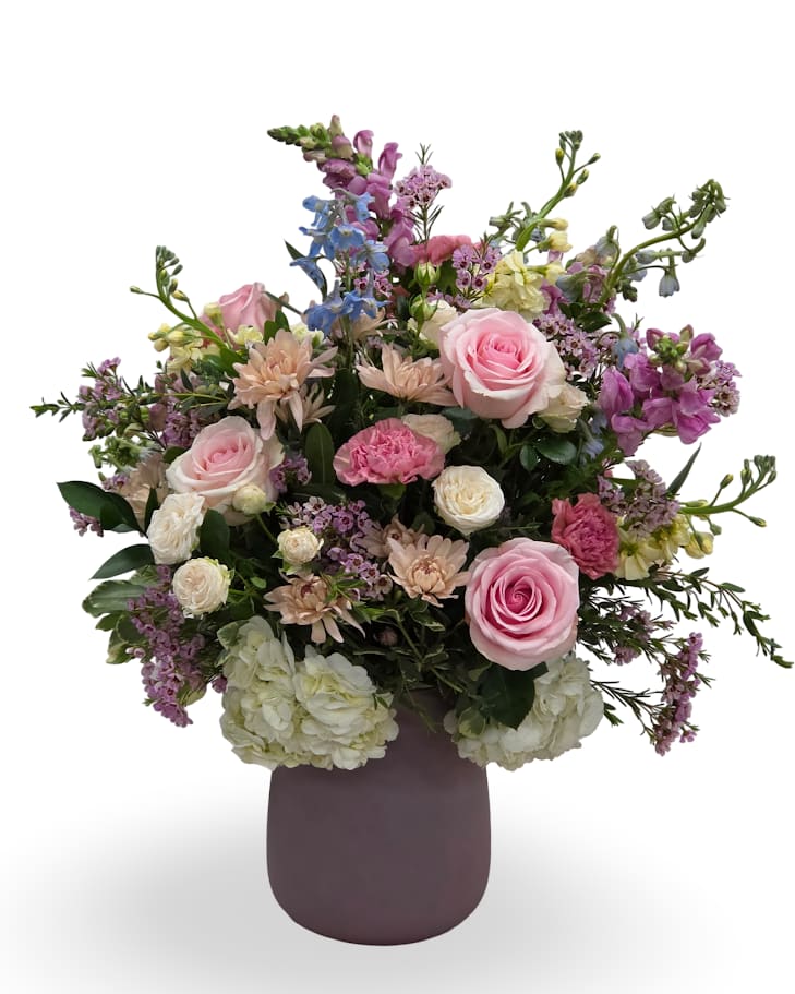 Blush Dream  - Soft blush, cream, lavender, and pale peach blooms spill in a lush, garden-style bouquet-ideal as an anniversary, birthday, or "just because" gift that feels effortlessly romantic. Designed to make the person you're thinking of feel celebrated and seen, this arrangement also brightens get-well wishes, thank-you moments, and just because moments.  Layered against dark green foliage and arranged with height and movement, the rounded composition comes in a matte pink ceramic vase that complements the soft color palette. Approximate dimensions are 26" x 23" x 21" inches, giving it a full presence on a table or mantle while still being easy to place in a room.