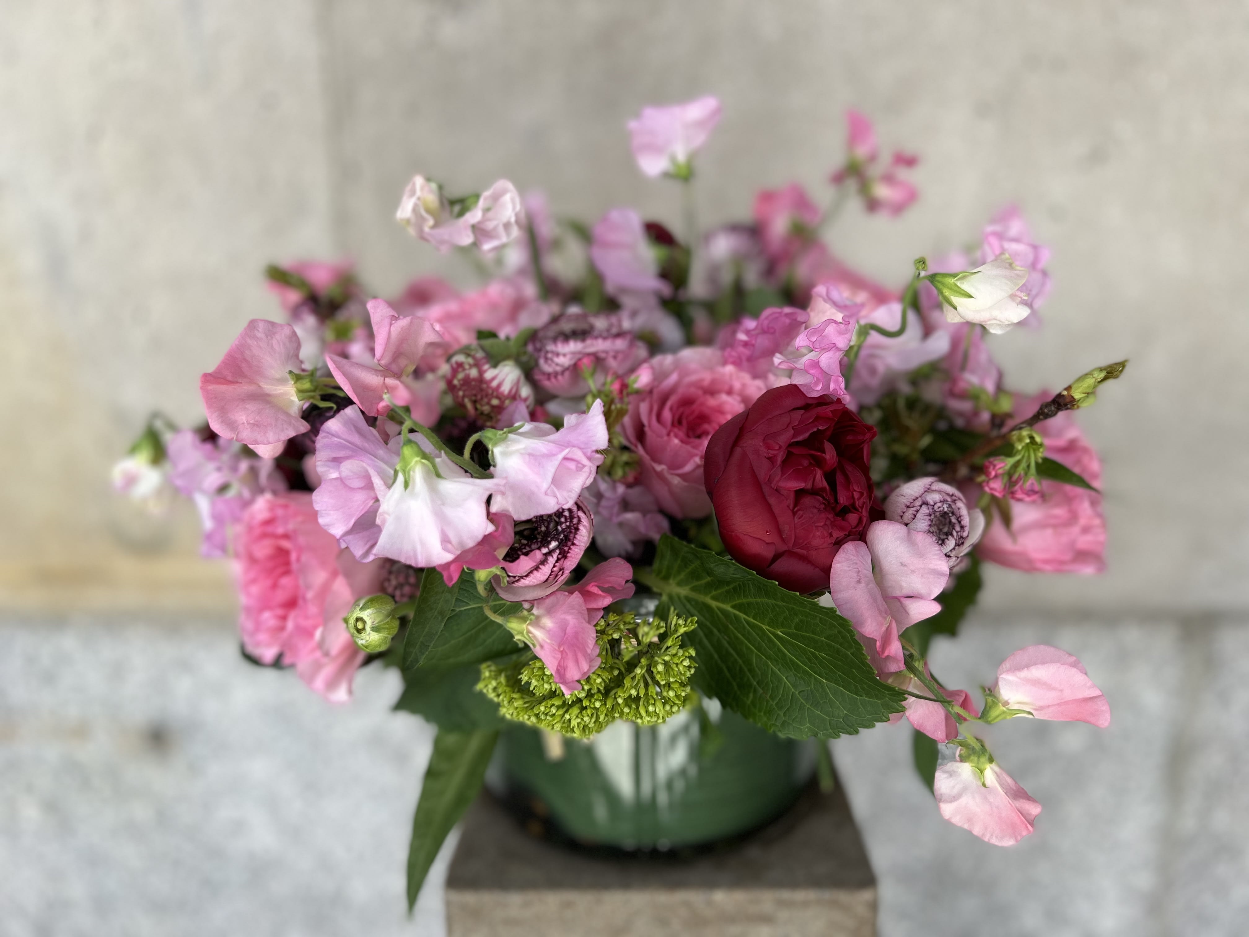 Strawberry Fields Forever  - This elegant arrangement, complemented by swirling garden roses , soft and subtle light pink sweet peas, white and pink asclepias and a touch of red charm peonies framed by lush hues of pinks colors and  textures like cappuccino ranunculos, ruffled greenery, and lofty stems of cherry blossoms in a clear round cylinder. 