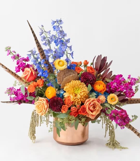 Autumn Joy - Embrace the enchanting hues of autumn with our "Autumn Joy" fall arrangement. This captivating display features striking blue delphinium, vibrant orange garden roses, rich orange ranunculus, and deep purple stock, all artfully arranged in a large copper pot. The combination of these bold colors and luxurious textures creates a warm, sophisticated centerpiece that beautifully captures the essence of a sunset in fall, perfect for adding a touch of elegance to your seasonal decor.  Our arrangements are unique, custom gifts designed by hand. Mother Nature changes our farm-fresh inventory every morning; to assure your gift is fresh and gorgeous our artists may need to make substitutions but will capture the color &amp; style of your selection to deliver a "WOW" for your recipient. 