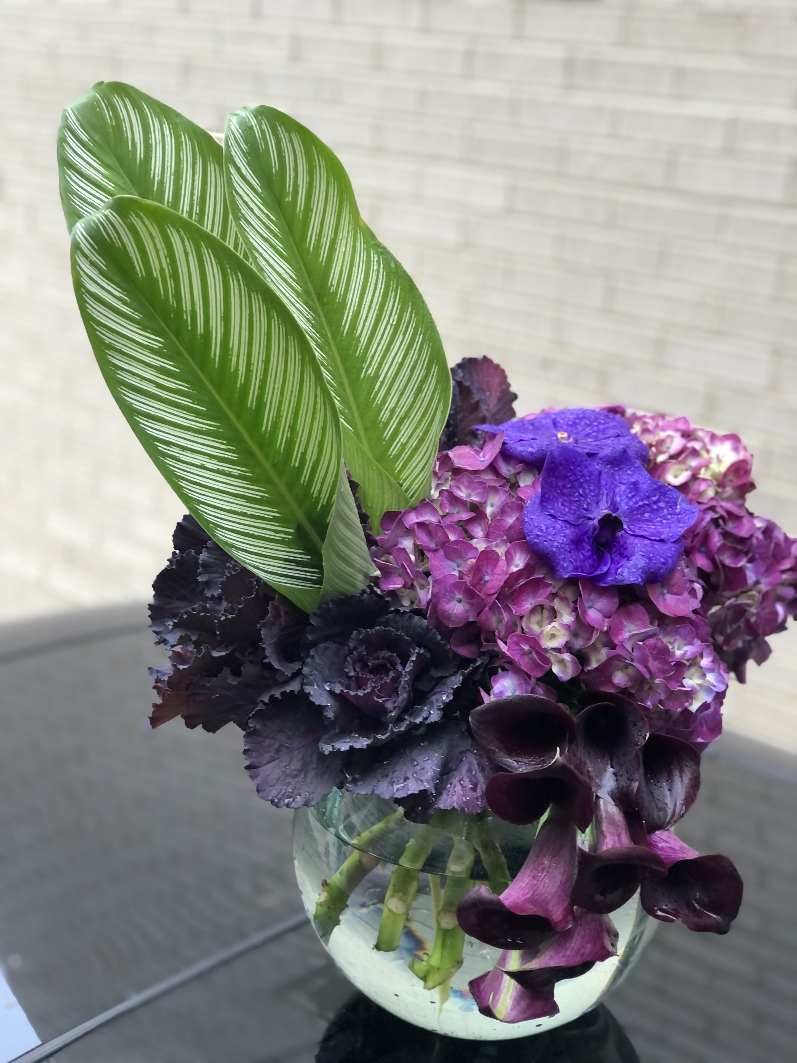 Purple hydrangea and dark calla lilies in a glass vase with striped green leaves