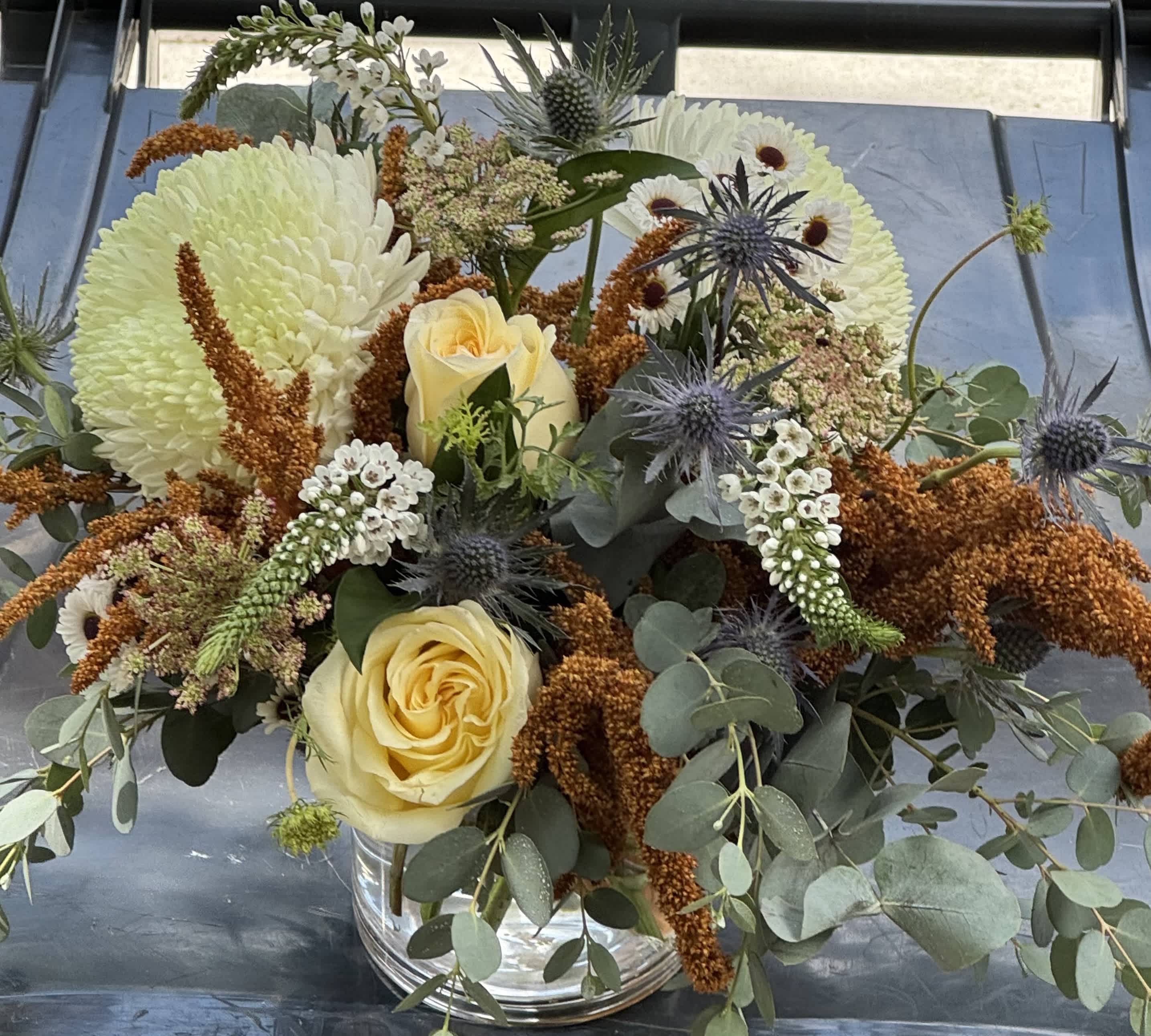 Bouquet of pale yellow roses, white chrysanthemums, and blue thistles in a glass vase