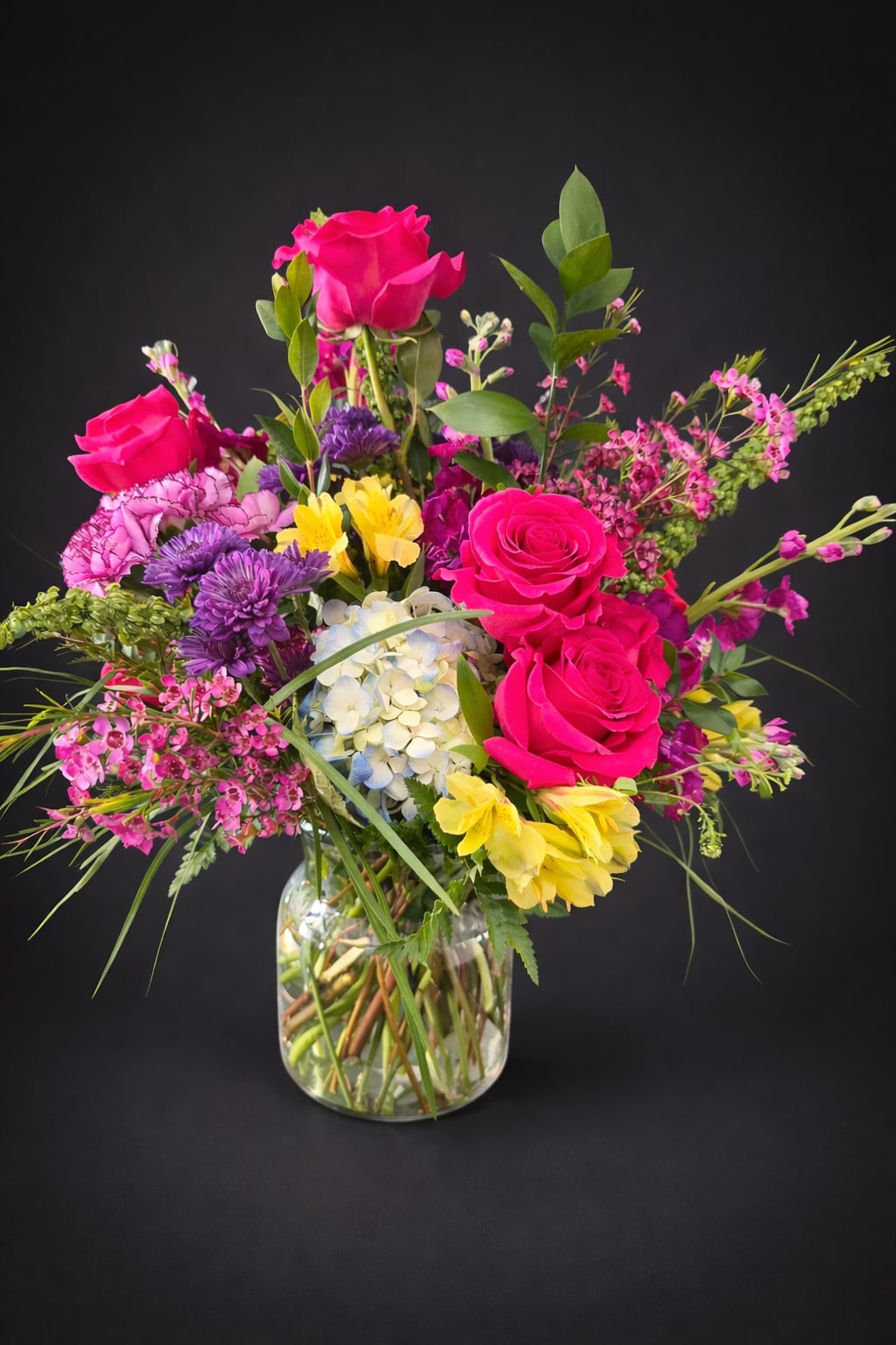 Garden Party Glow - This Colorful Rose and Hydrangea Vase Arrangement features hot pink roses, blue hydrangea, yellow alstroemeria, purple mums, and pink seasonal accents in a clear glass vase. This bright mixed flower arrangement is perfect for birthdays, celebrations, thank-you gifts, and everyday flower delivery. Designed in a gathered garden style, it brings fresh color, texture, and charm to any occasion.