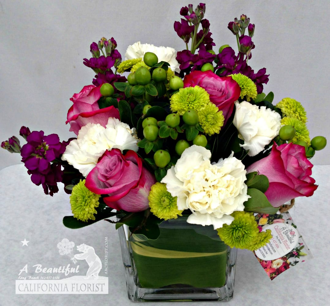 Pink roses, white carnations, and purple stock in a square glass vase