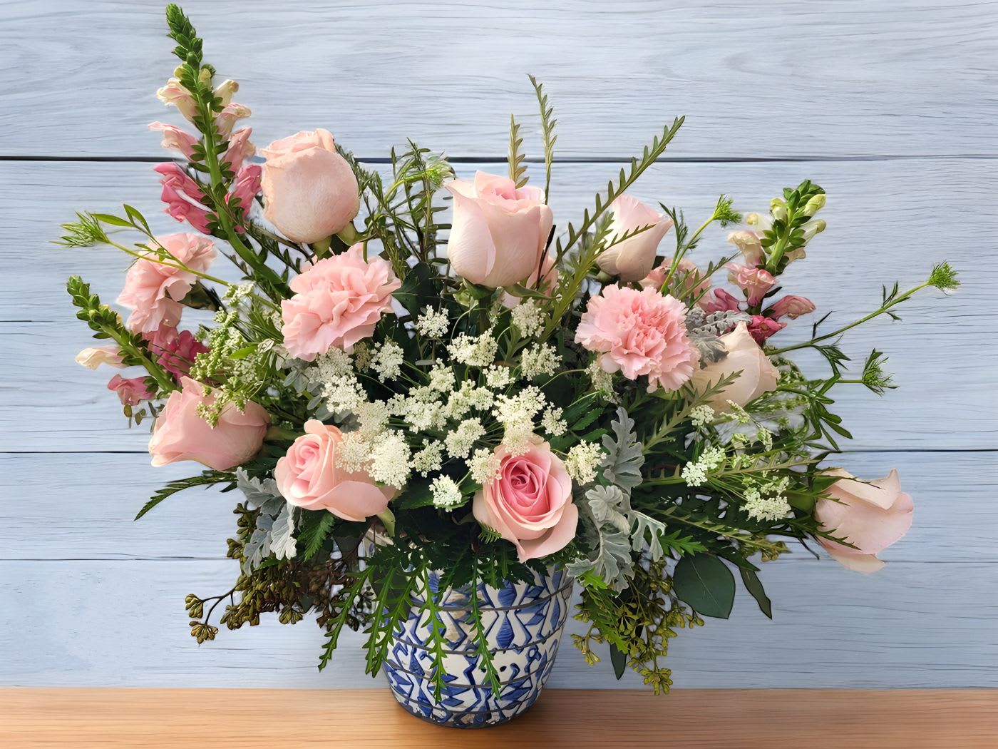 Blushing Beauty - Show someone you care with the 'Blushing Beauty' a stunning arrangement that is sure to delight. The soft hues of light pink roses, carnations, and snapdragon create a truly captivating display. Accented with Queen Anne's Lace, dusty miller, and lush greens, this arrangement is sure to bring joy and brighten any room. Presented in a beautiful 6" diameter blue and white ceramic vase, it's a thoughtful gift for any occasion.  Approximate size: 18"Tall x 24" Wide Orientation: All Around